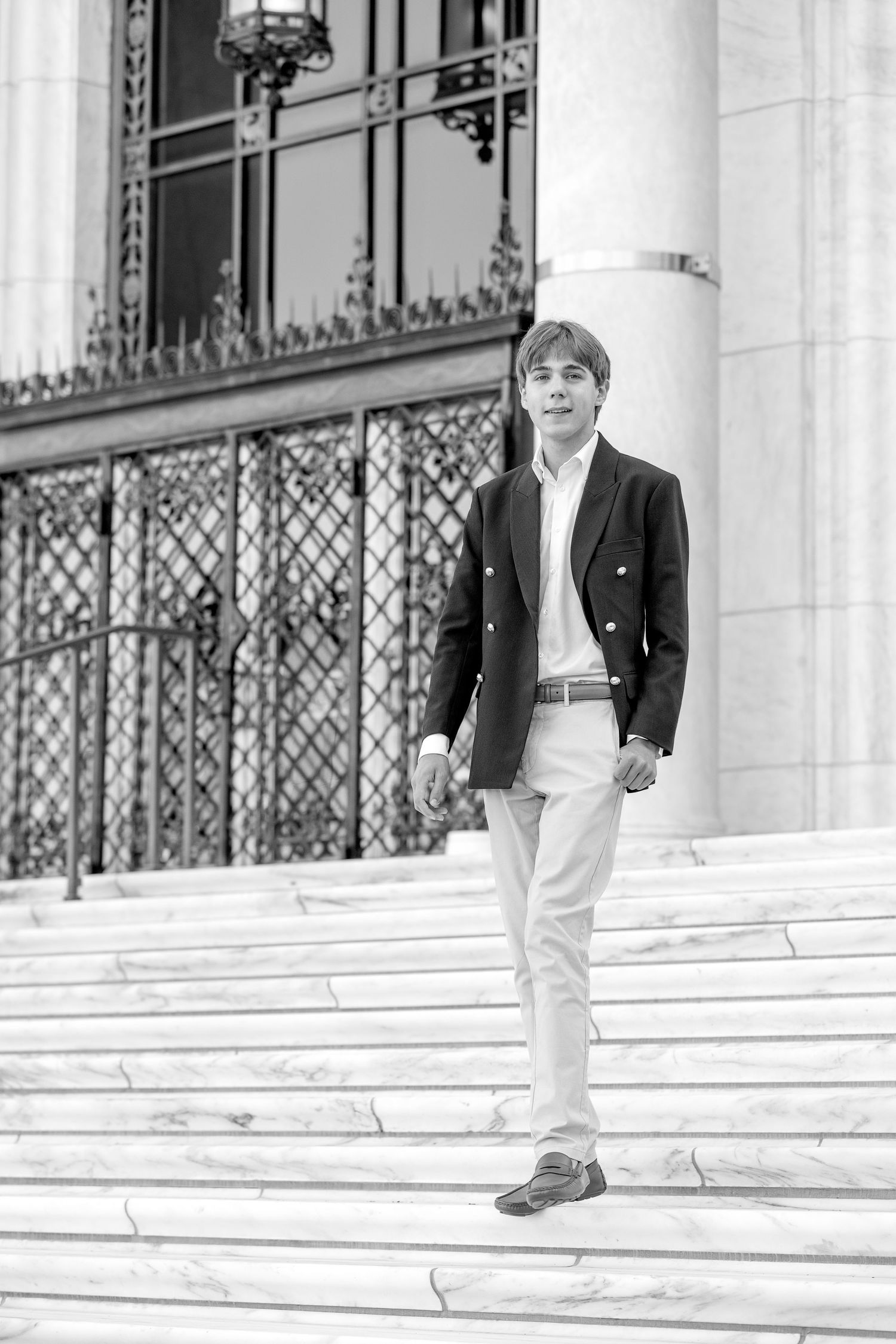 Young man in blazer and light pants descending marble steps of elegant building in black and white photo.