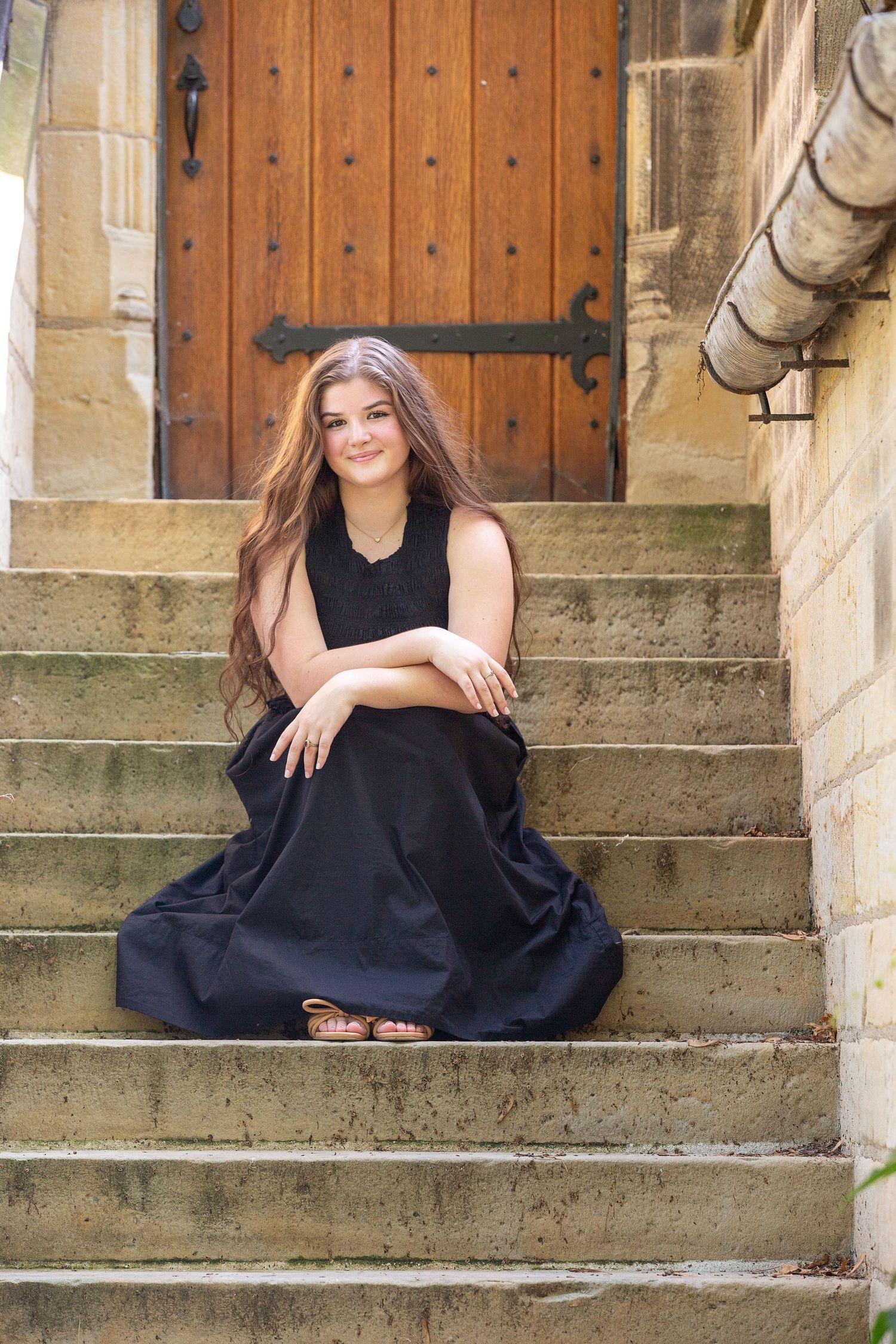 Young woman in black maxi dress sitting on stone steps near ornate wooden door, smiling.