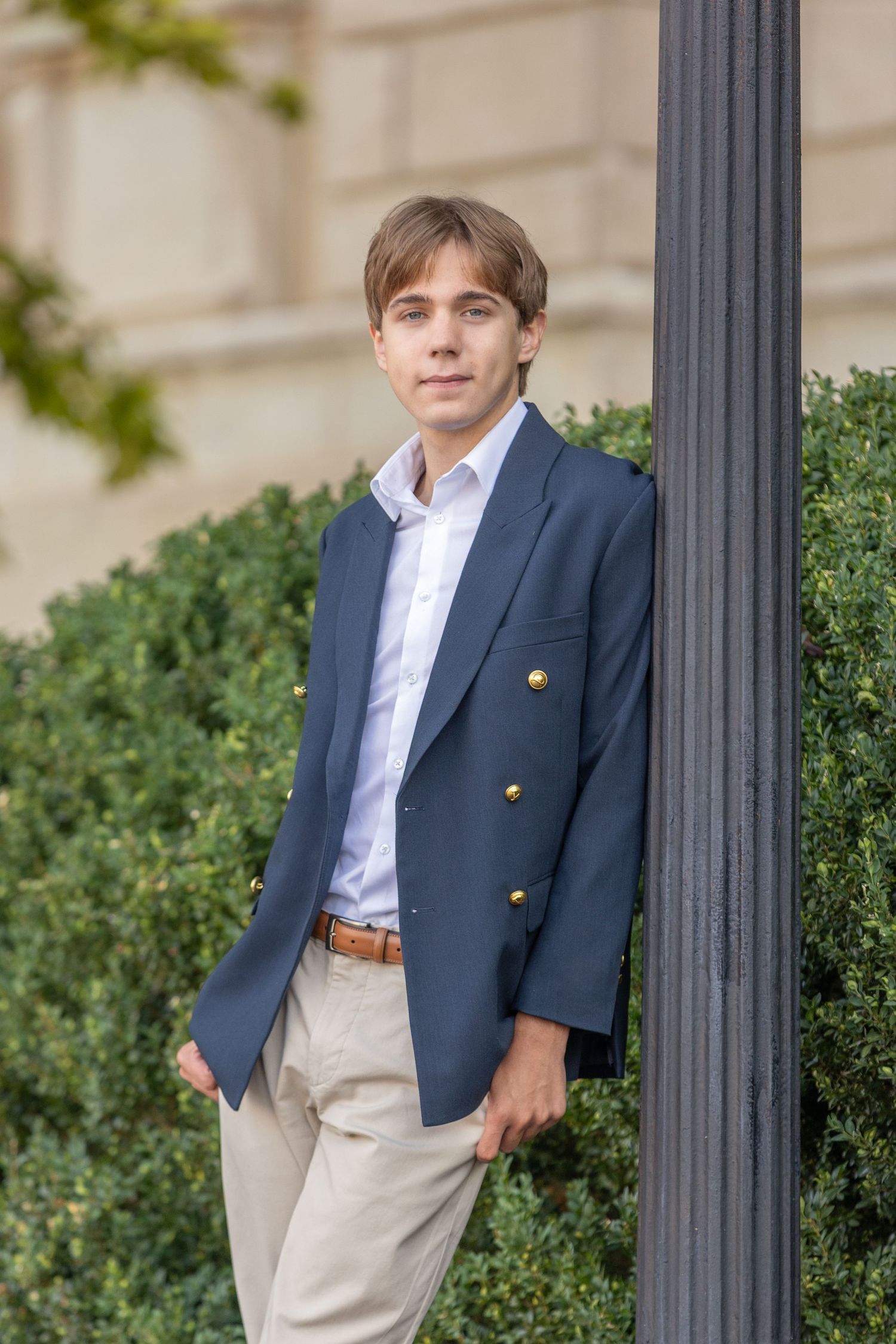 Young man in navy blazer with gold buttons and khaki pants leaning against a lamppost outdoors.