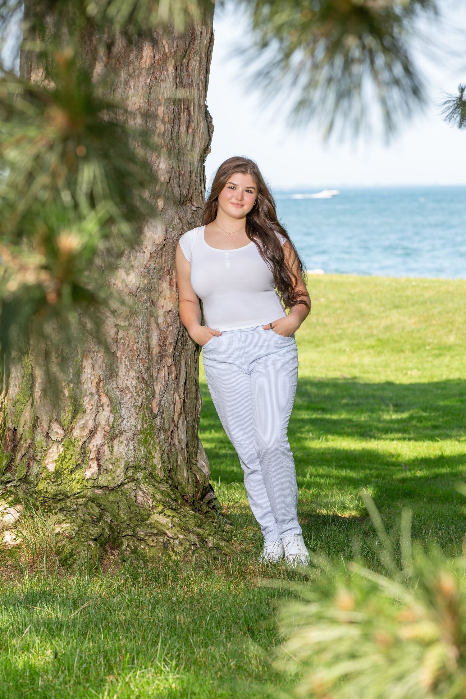 Young woman in white top and jeans leaning against a tree in a park with ocean view behind her.