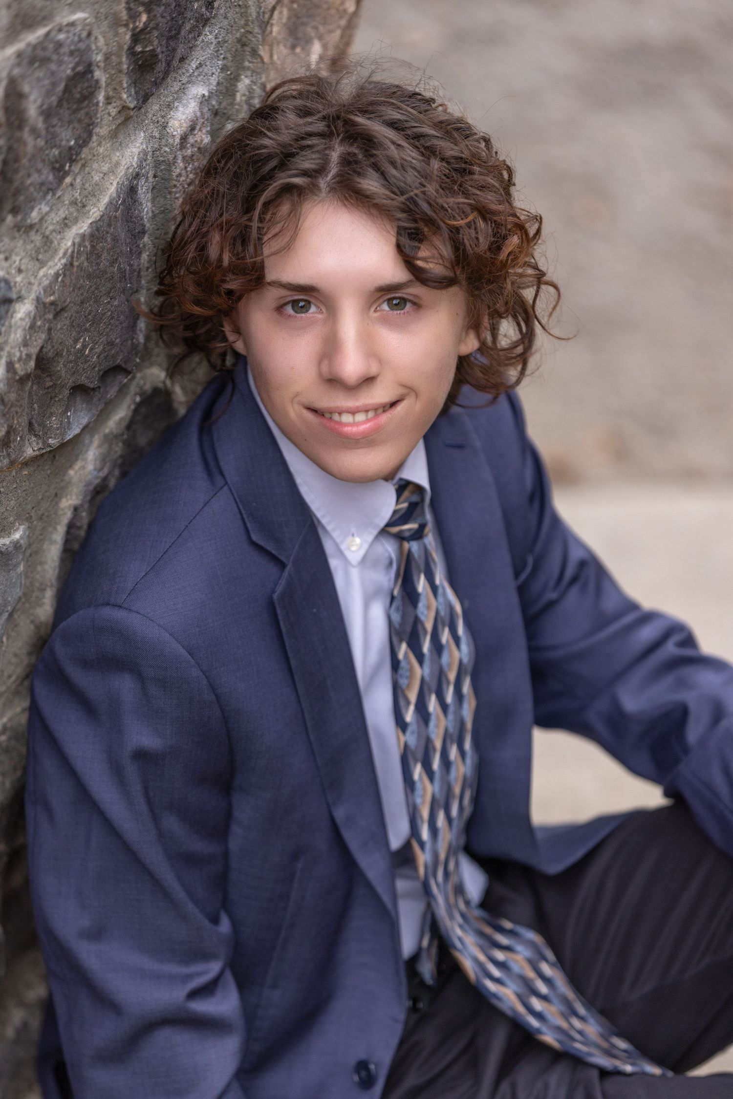 Young man with curly hair in a blue suit and patterned tie, leaning against a stone wall outdoors.