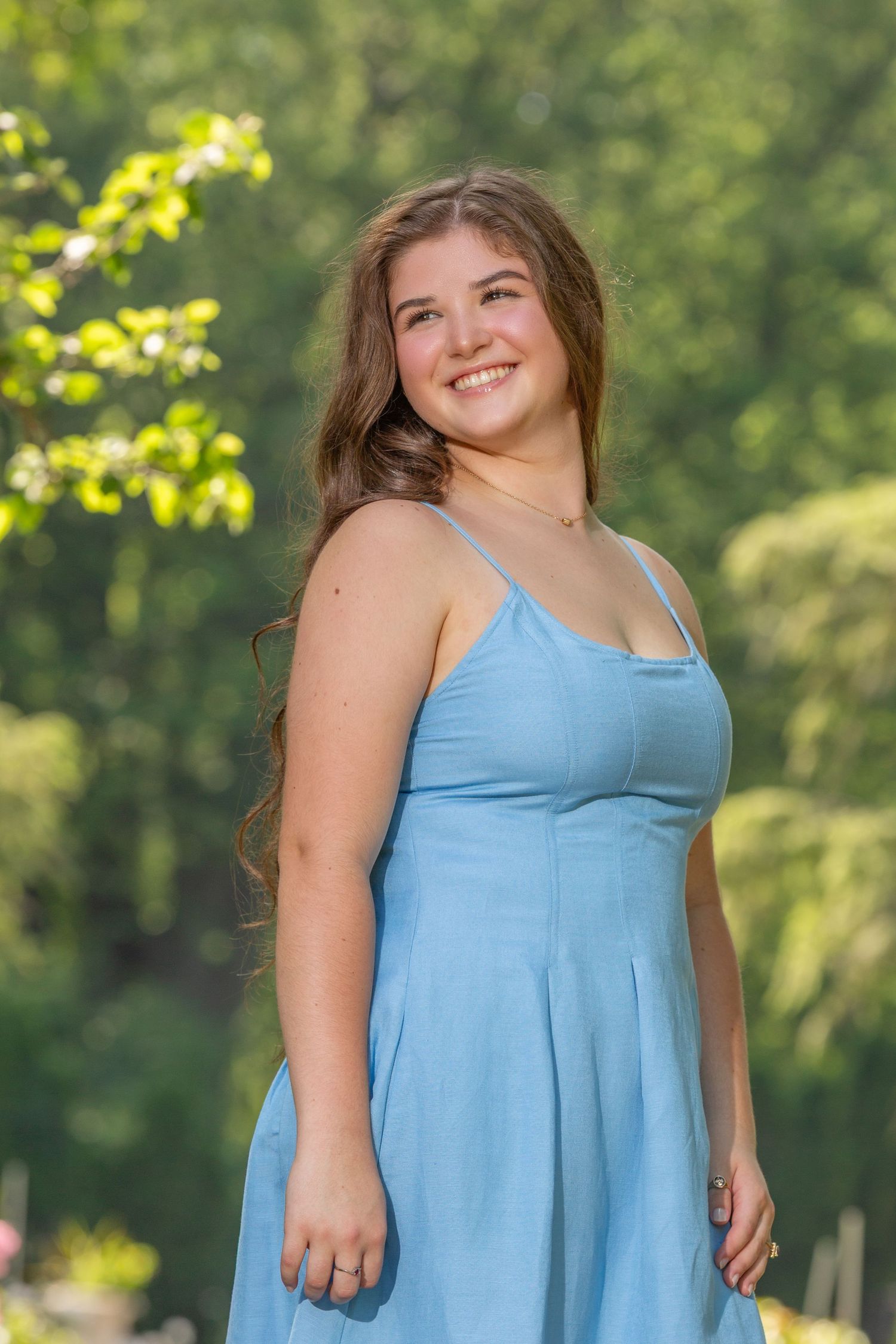 Smiling young woman in a light blue sundress posing outdoors with lush green trees in the background.