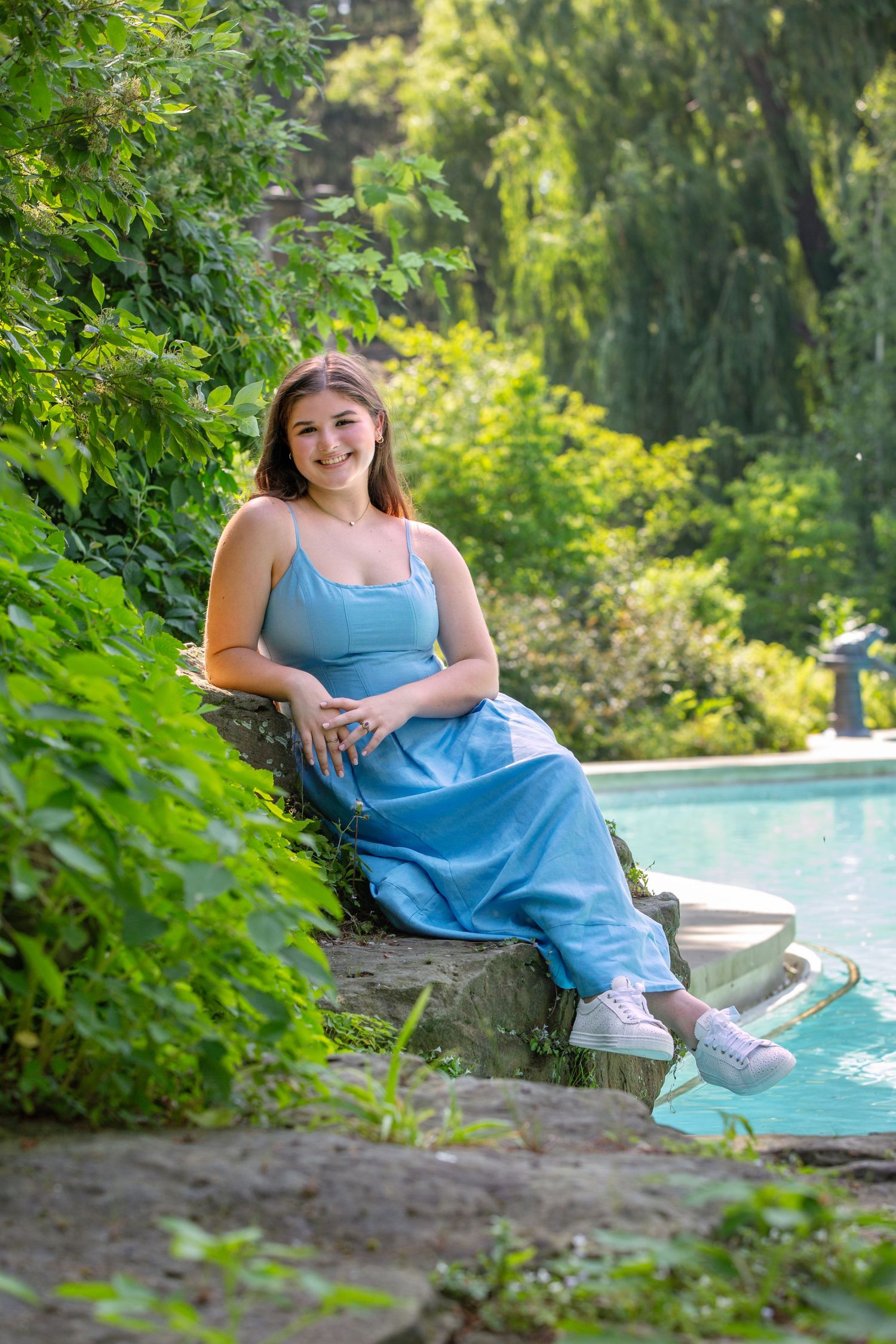 Young woman in blue dress sits on stone wall by pool surrounded by lush green garden, smiling warmly.
