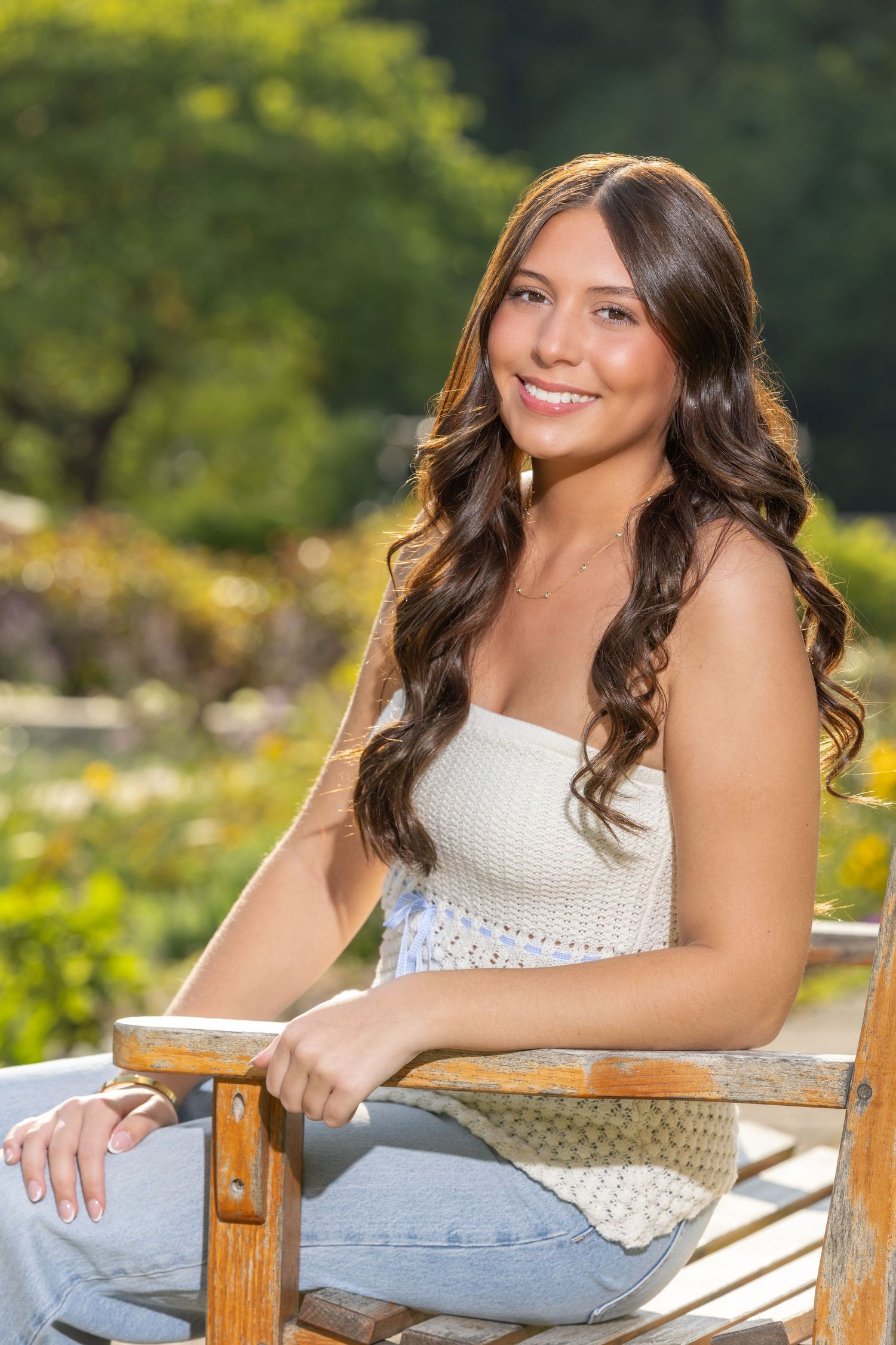 Young woman with long curly hair smiling outdoors, sitting in wooden chair wearing white crop top and jeans.