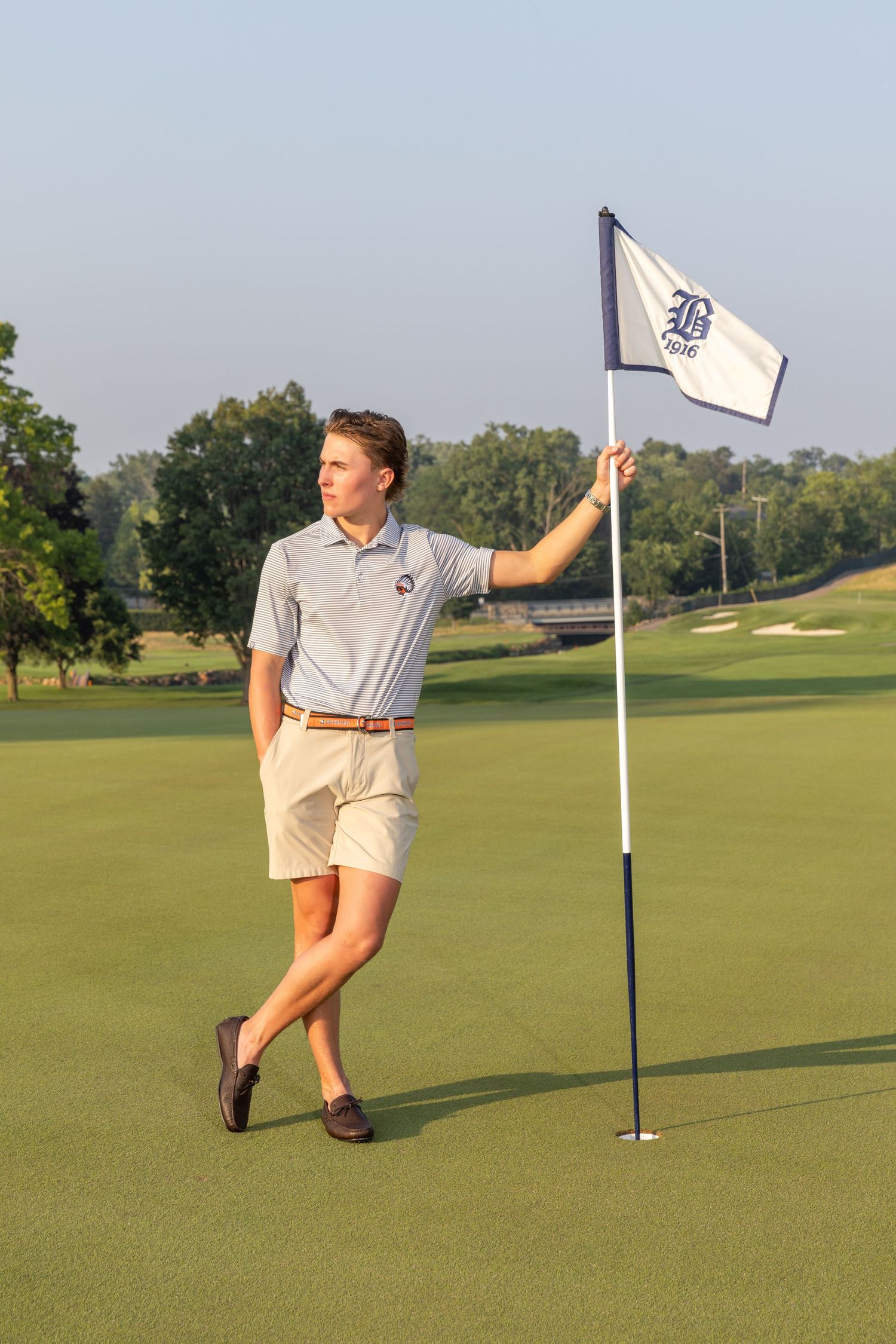 Young man in polo shirt and khaki shorts holding a golf flag on a lush green golf course on a sunny day.