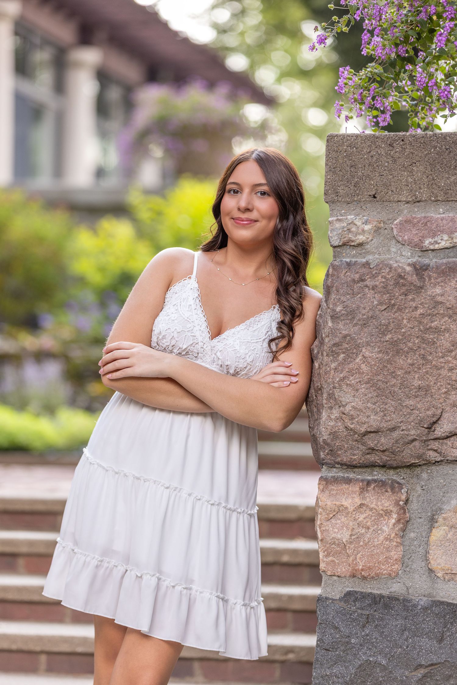 Young woman in white sundress leaning against stone pillar outdoors, smiling with arms crossed, purple flowers above.