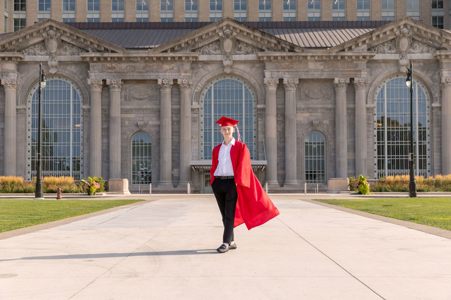 Graduate in red cap and gown walks proudly in front of a grand university building on graduation day.