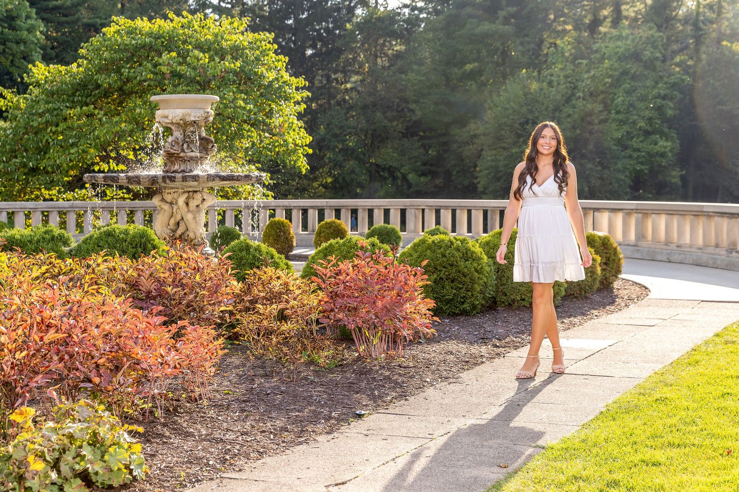 Young woman in white dress walking along garden path surrounded by colorful shrubs.