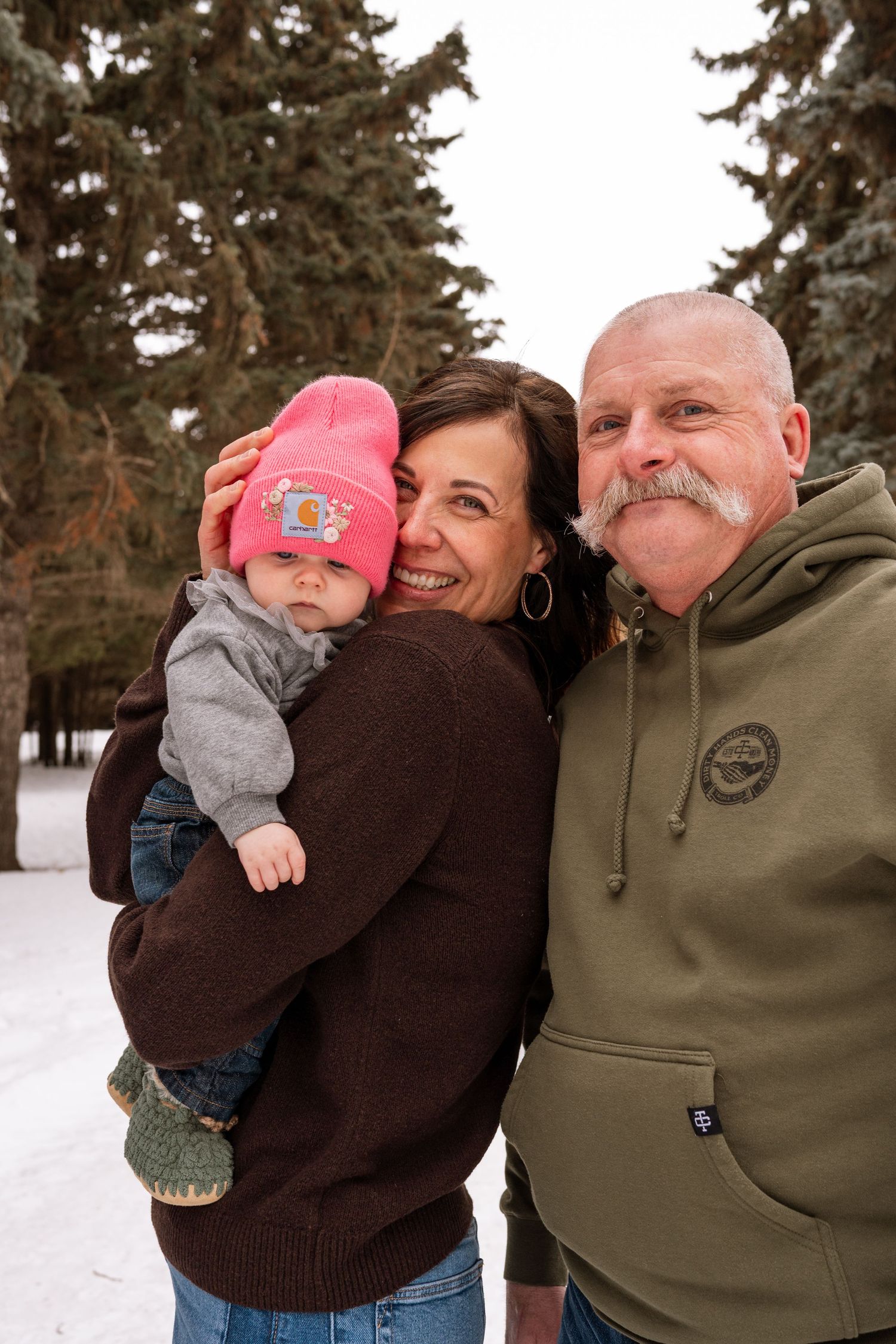 Grandparents smiling outdoors in winter, holding a baby in a pink hat, surrounded by snow-covered evergreen trees.
