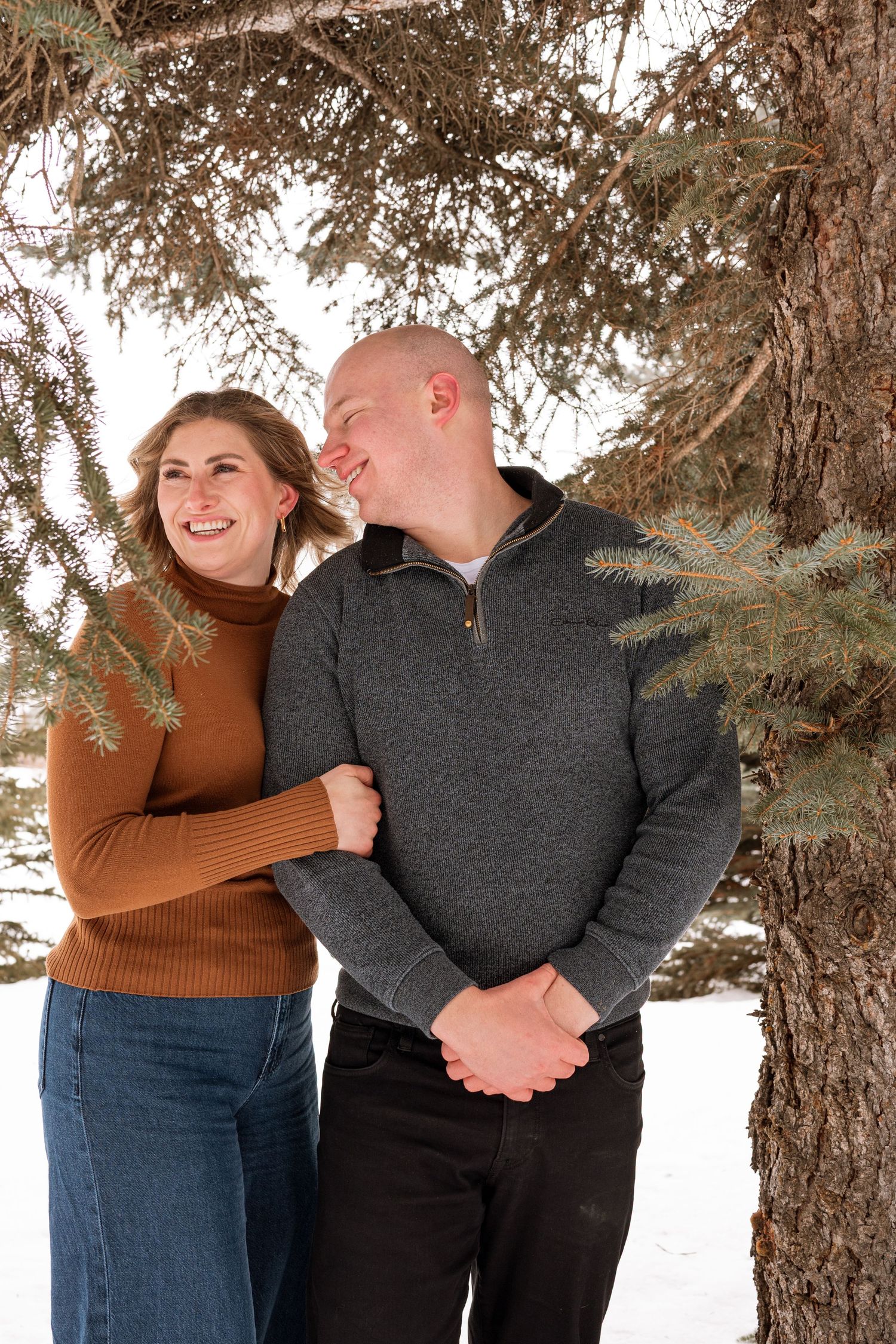 Couple embracing and laughing under pine tree branches in snowy winter setting, woman in brown turtleneck and man in gray sweater.