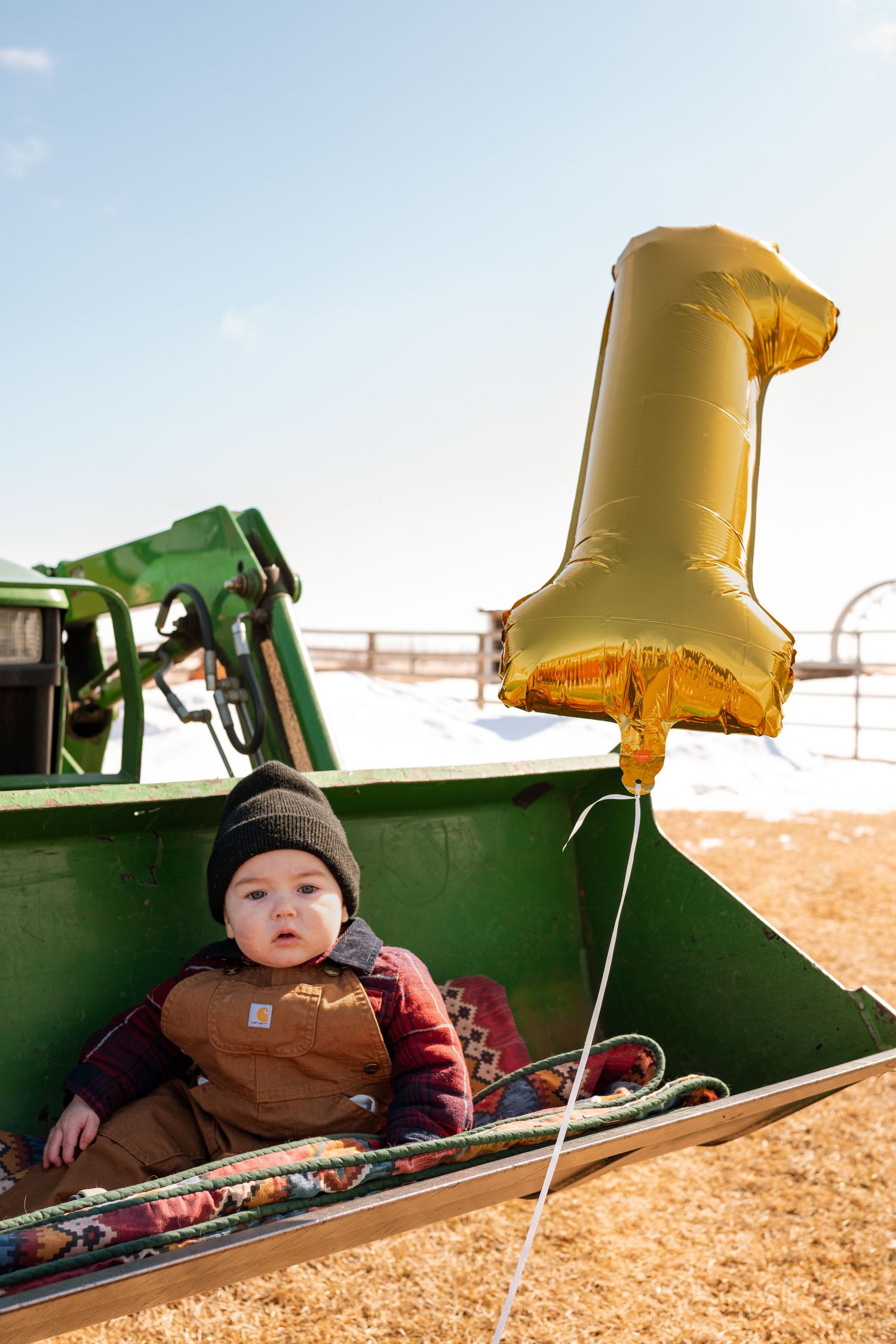 Baby in Carhartt overalls sits in green tractor bucket with gold number 1 balloon celebrating first birthday outdoors.