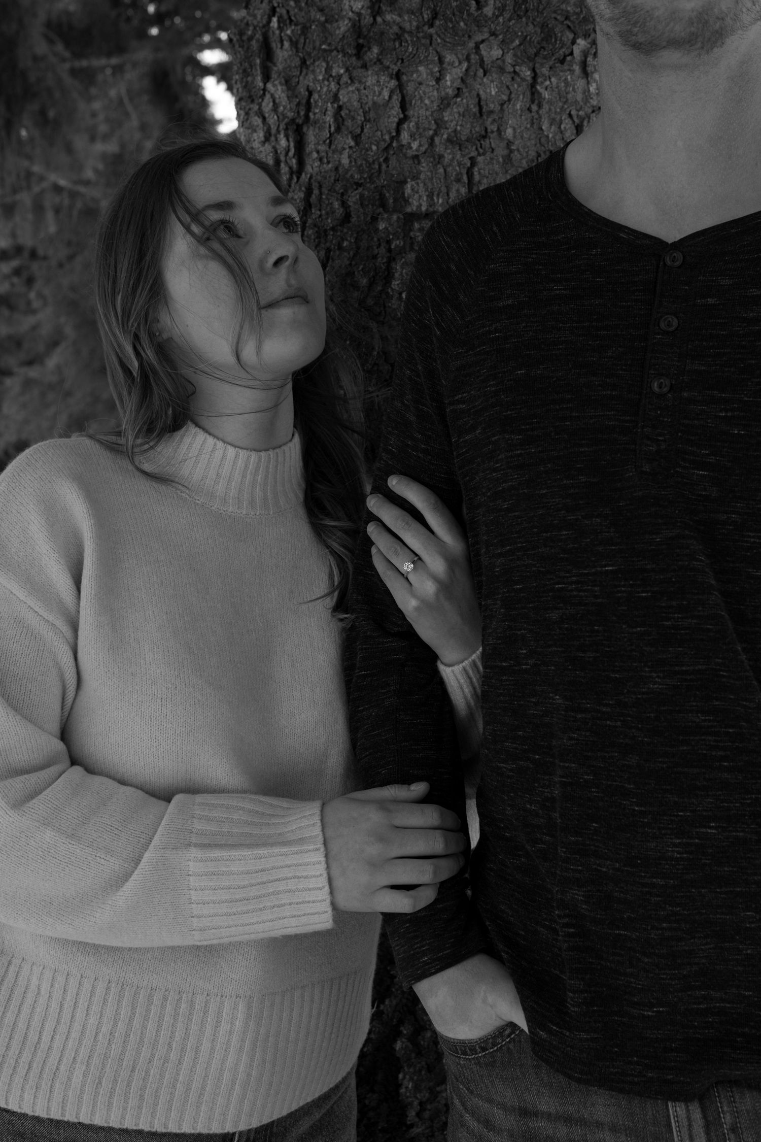A woman in a cream turtleneck sweater leans against a tree beside a man in a black henley in black and white.
