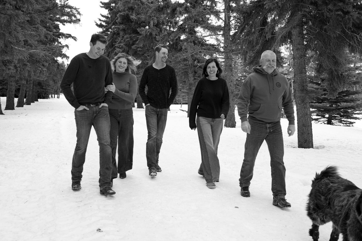 Family of five walking together with a dog on a snowy path surrounded by evergreen trees in winter.