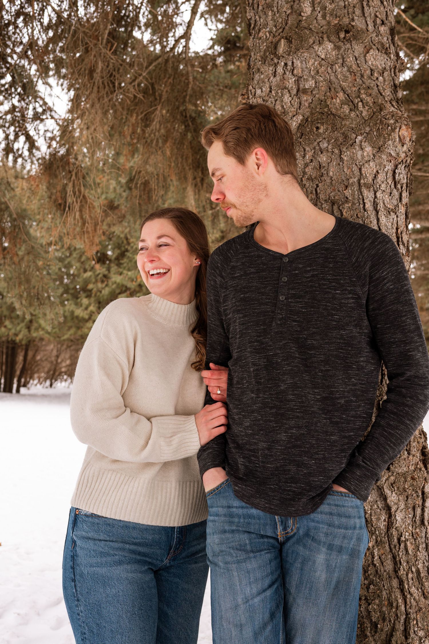Happy couple laughing together leaning against a tree in a snowy winter park setting.