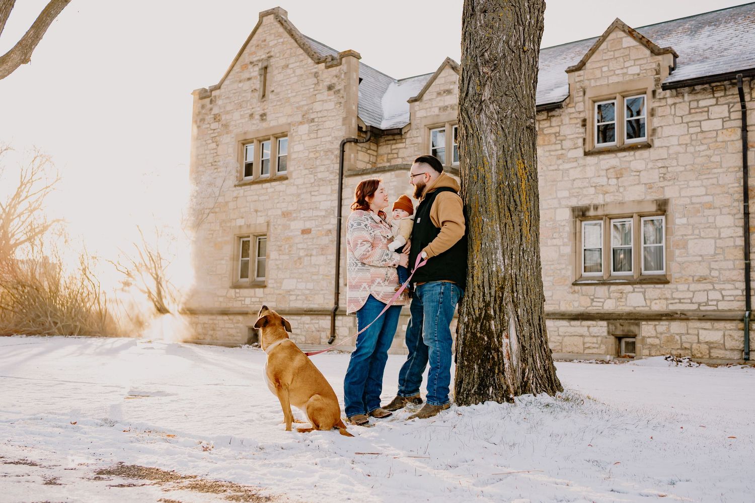 A couple and their dog stand in the snow outside stone buildings during winter.