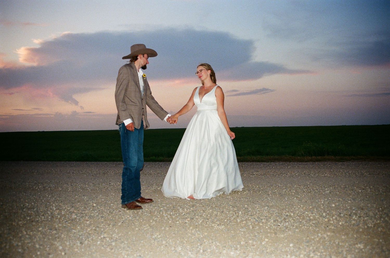 A couple in western wedding attire holds hands on a dirt road at sunset with a dramatic cloudy sky.