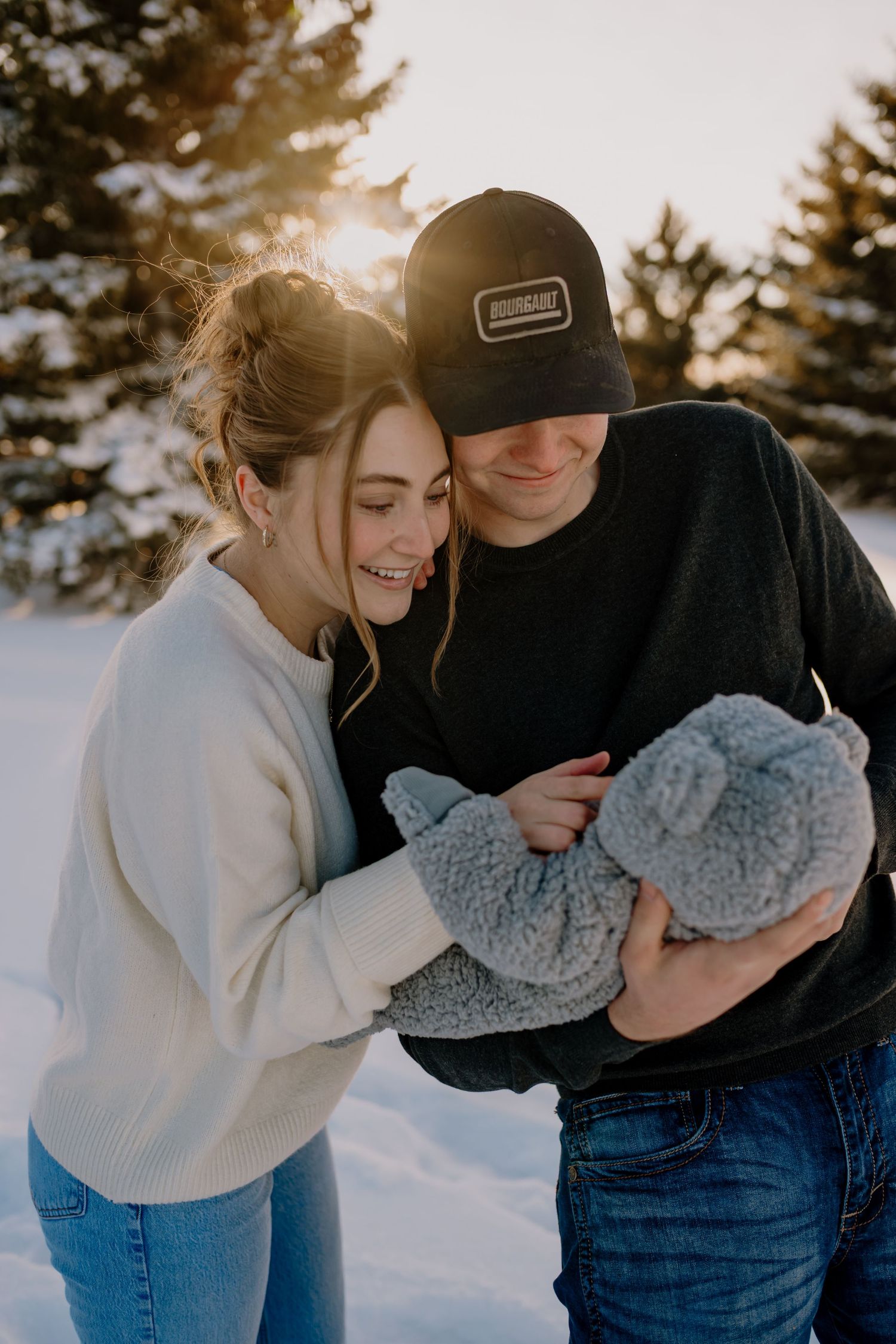 A couple shares a tender moment holding a baby in winter, with sunlight peeking through snow-covered trees behind them.