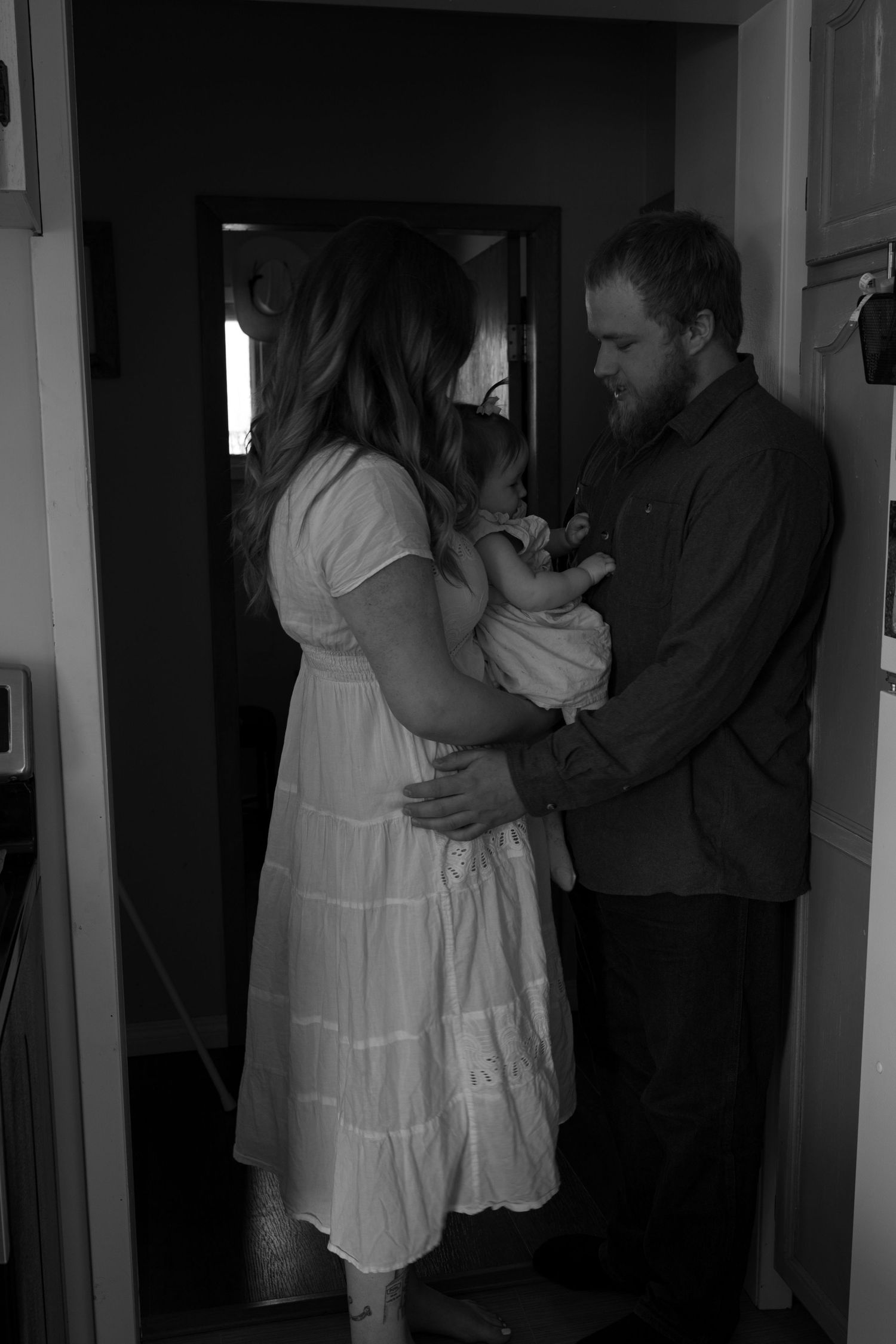 Black and white photo of a couple in a doorway tenderly holding a newborn baby together.