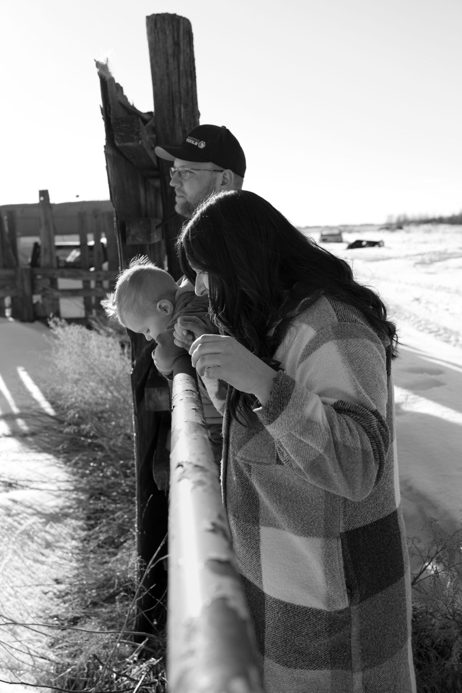 Black and white photo of a couple with baby at a rustic wooden fence on a snowy rural road.