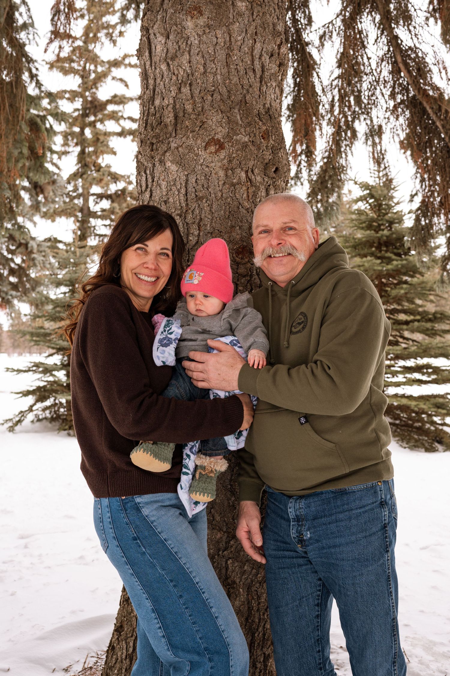 Smiling grandparents holding a baby in a pink knit hat while standing by a large tree in a snowy park.