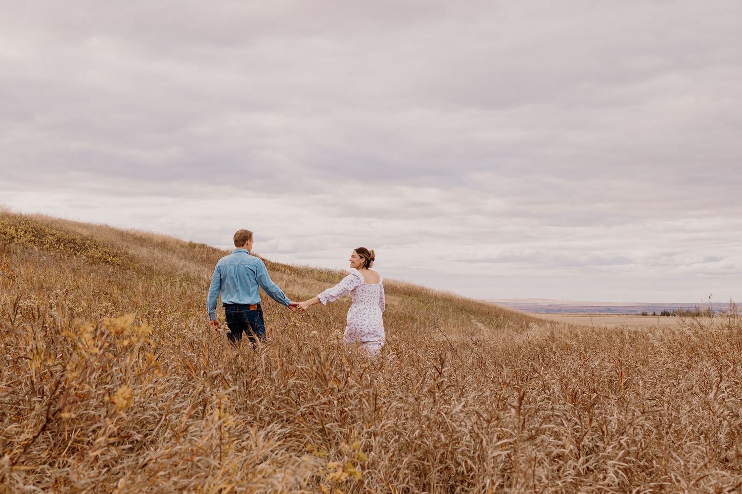 A couple walks through a golden wheat field on an overcast day with rolling hills in the background.