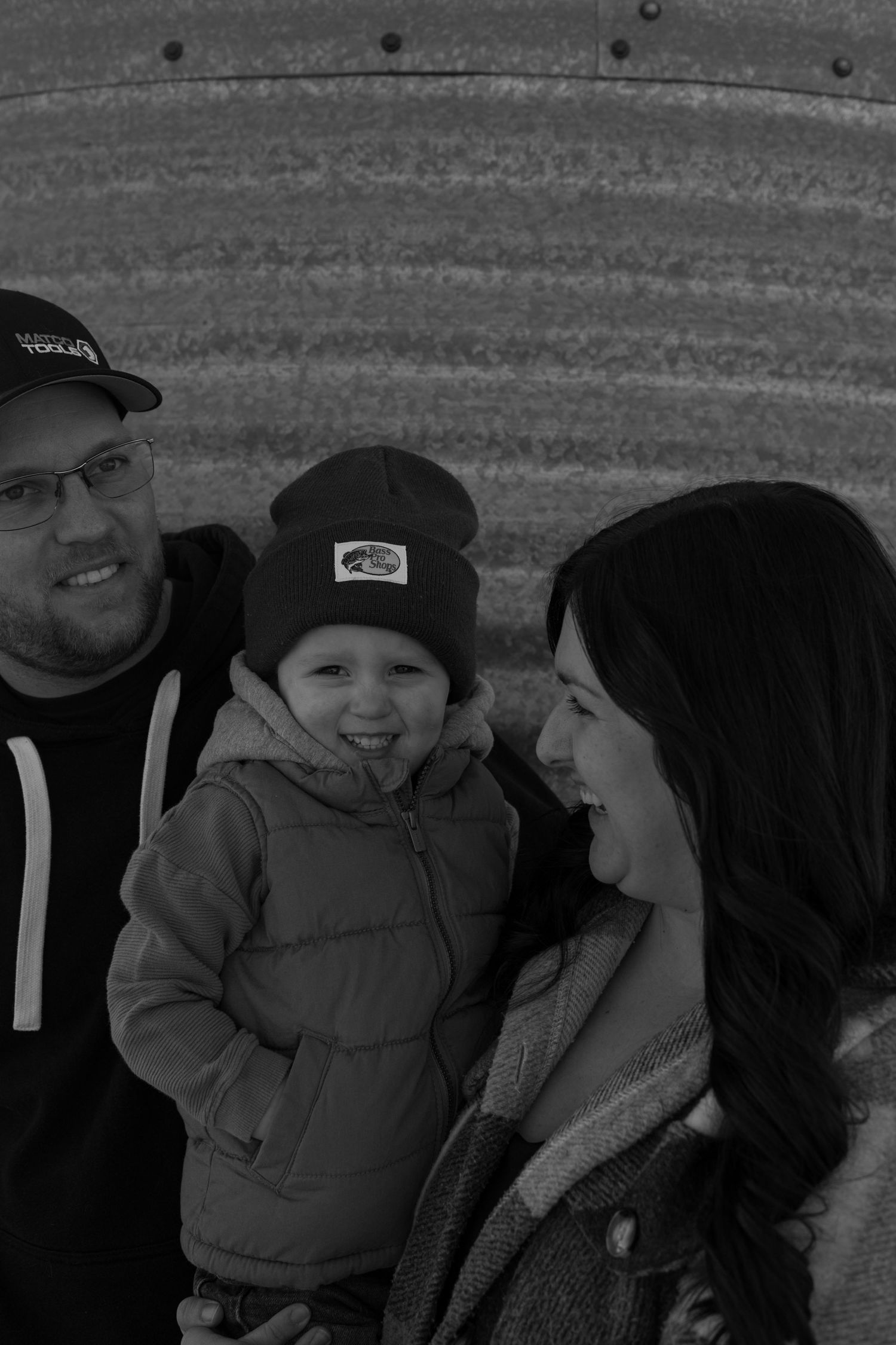 Black and white photo of a smiling toddler in a beanie hat held by two adults near a corrugated metal wall.