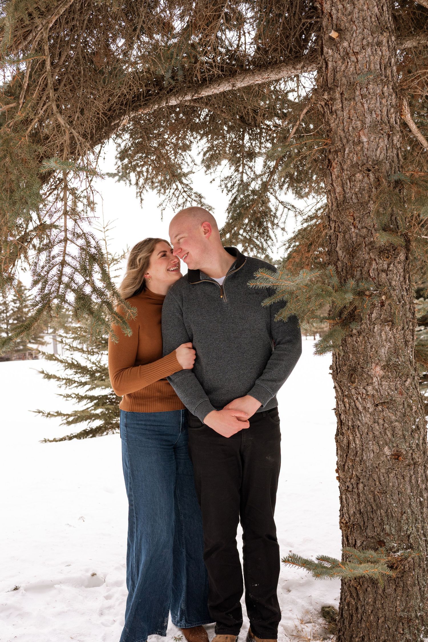 A couple shares a romantic moment under snow-dusted pine trees in a winter landscape, smiling at each other warmly.