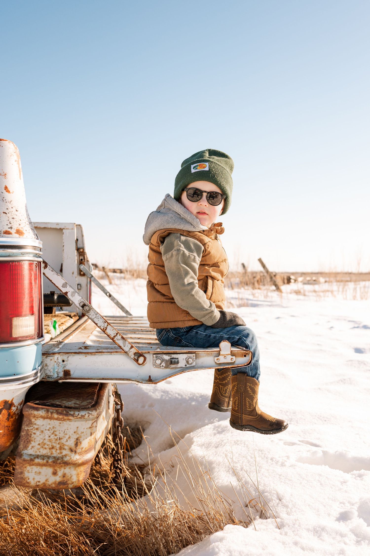 Stylish young boy in sunglasses and green beanie sits on vintage snowy truck bumper.