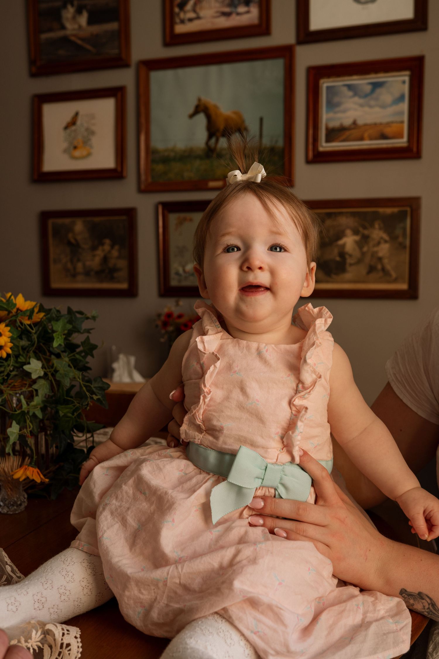 Smiling baby girl in pink dress with mint bow sits happily surrounded by framed artwork on wall.