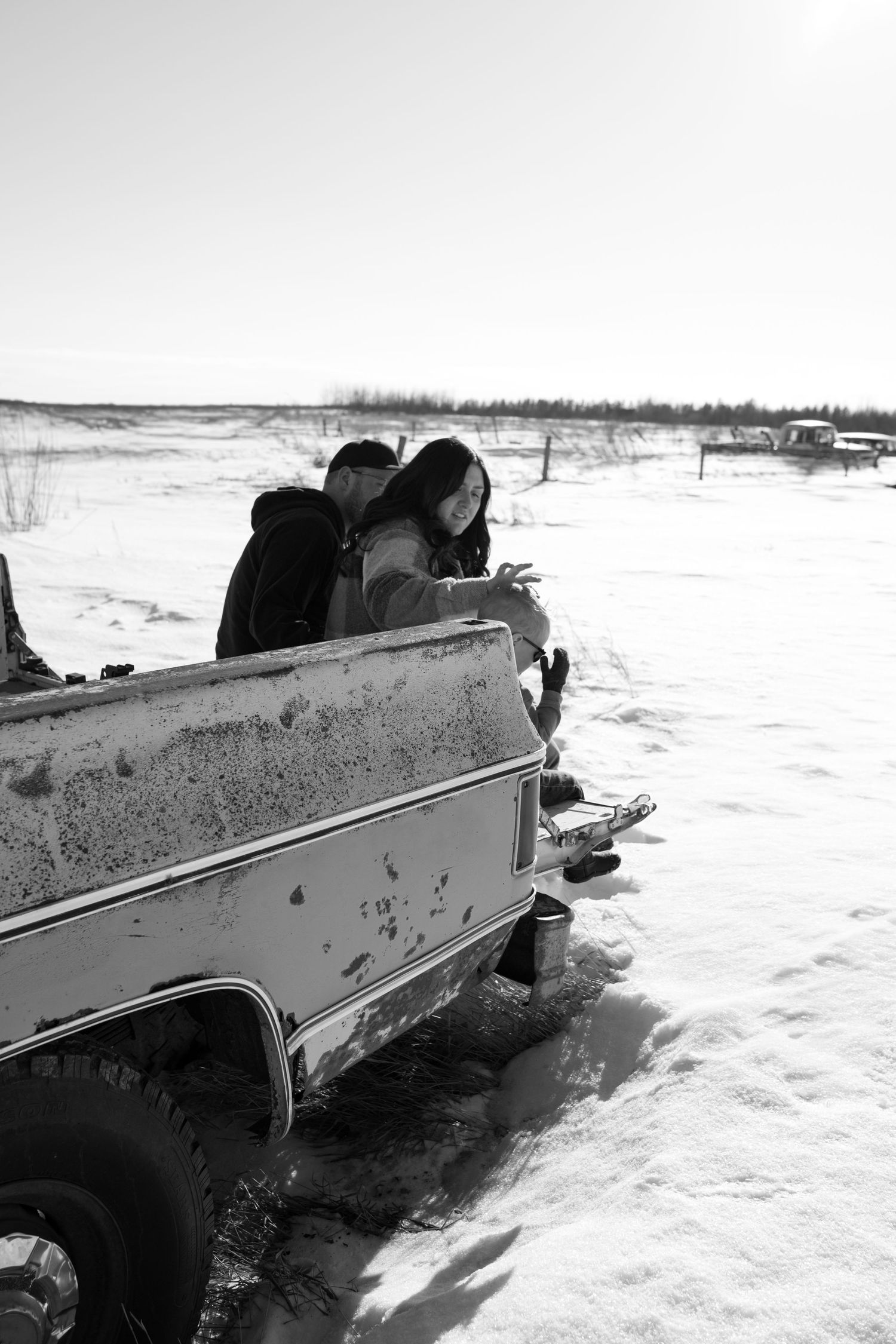Black and white photo of couple leaning over snowy vintage truck bed in winter landscape.