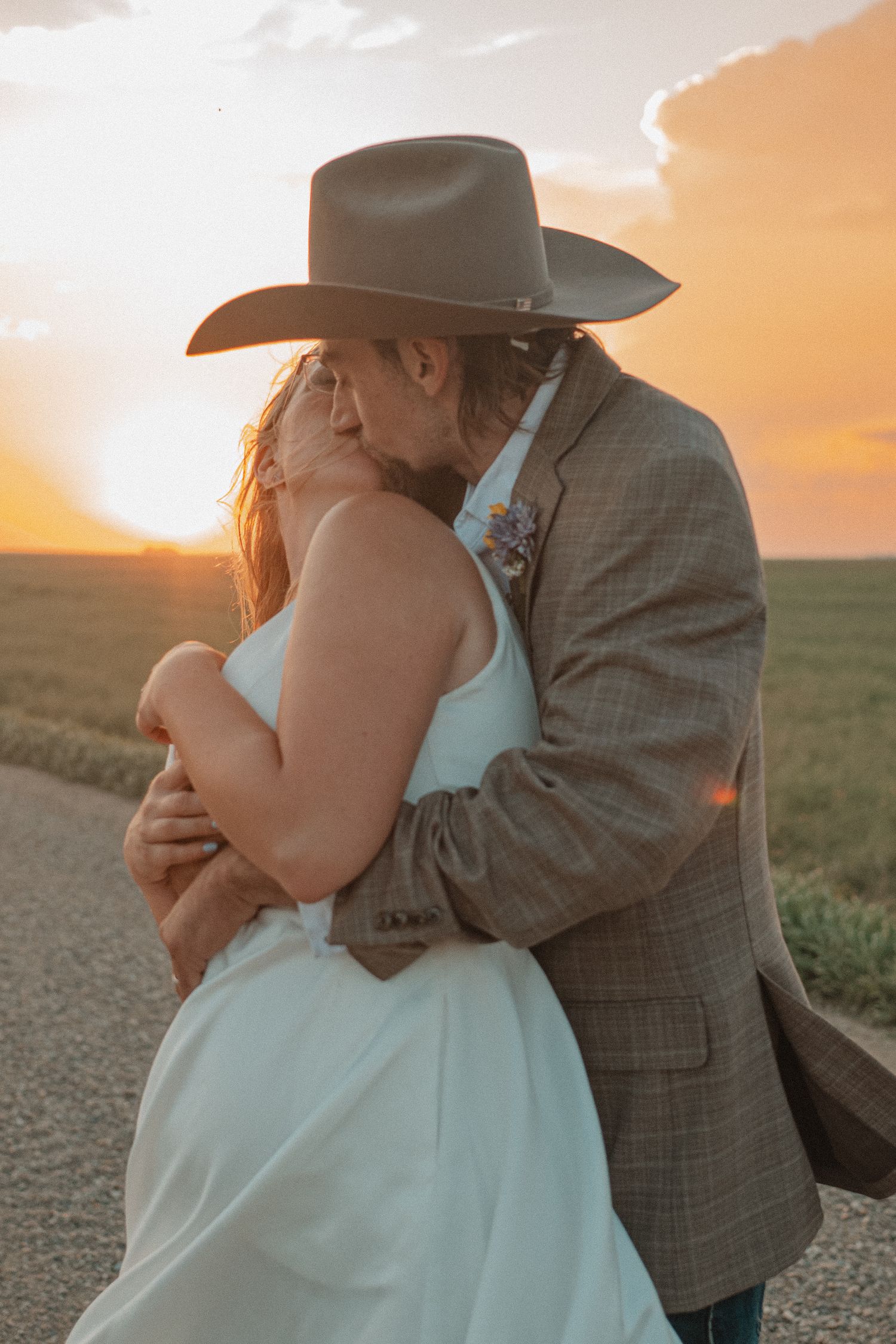 A romantic couple embraces during sunset in a rural field while wearing western attire and formal dress.