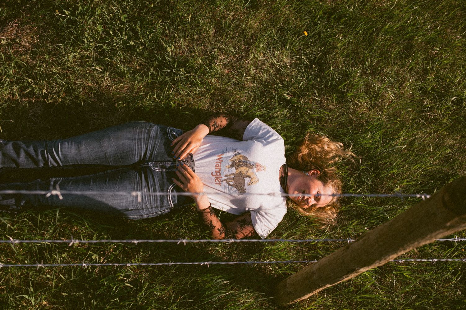 Aerial view of a person lying on green grass wearing jeans and a white t-shirt near a wire fence.