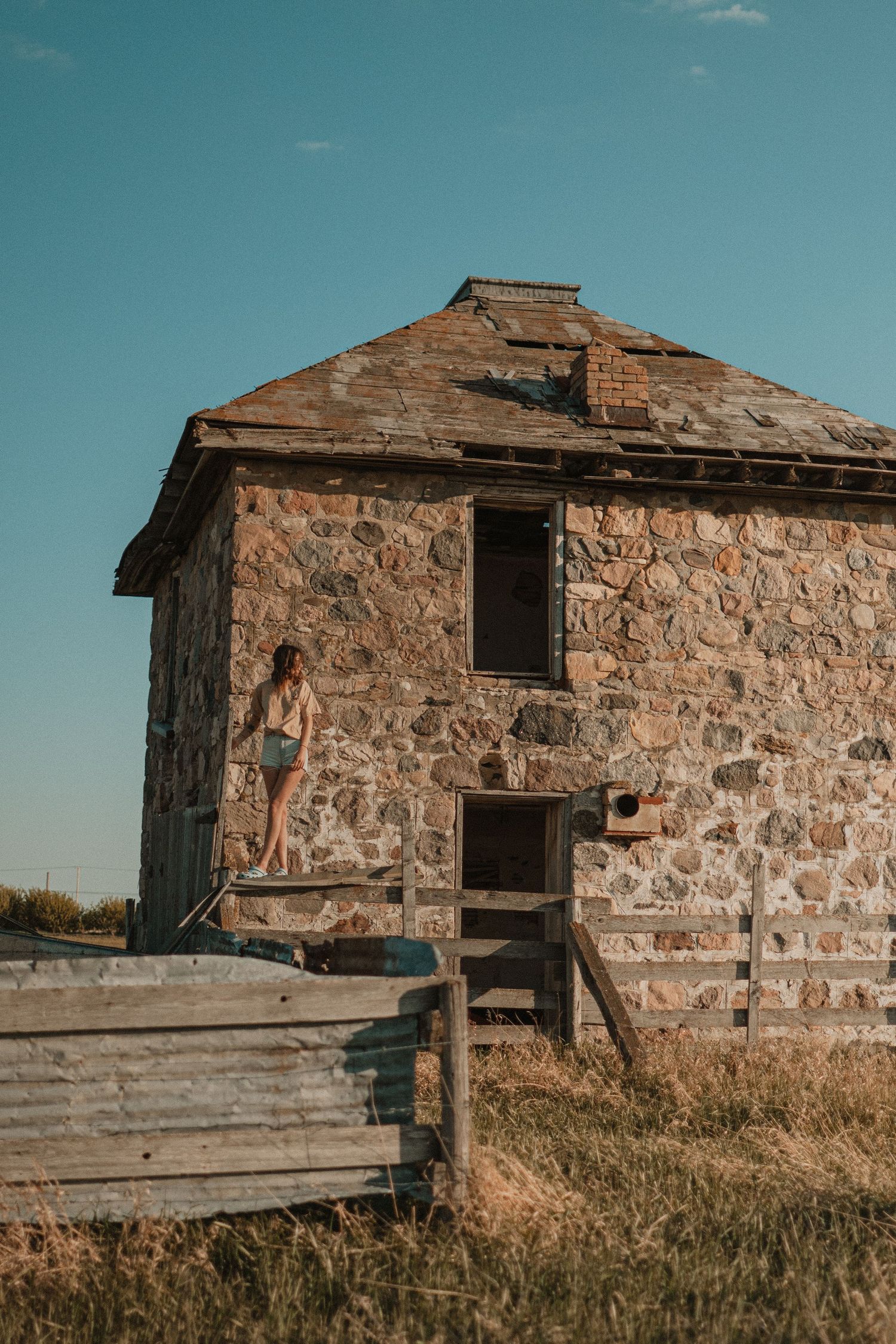 A vintage stone house with wooden steps and rustic architectural details stands against a turquoise sky in a rural setting.