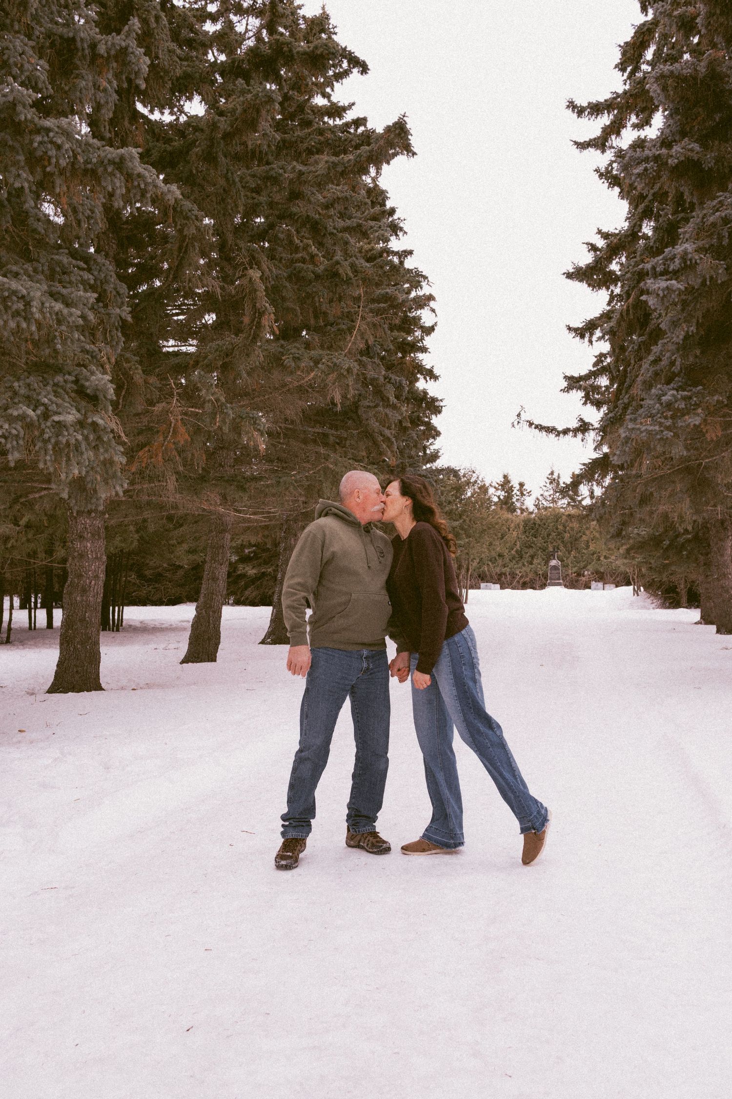 Couple sharing a kiss on a snowy path lined with tall evergreen trees in winter.