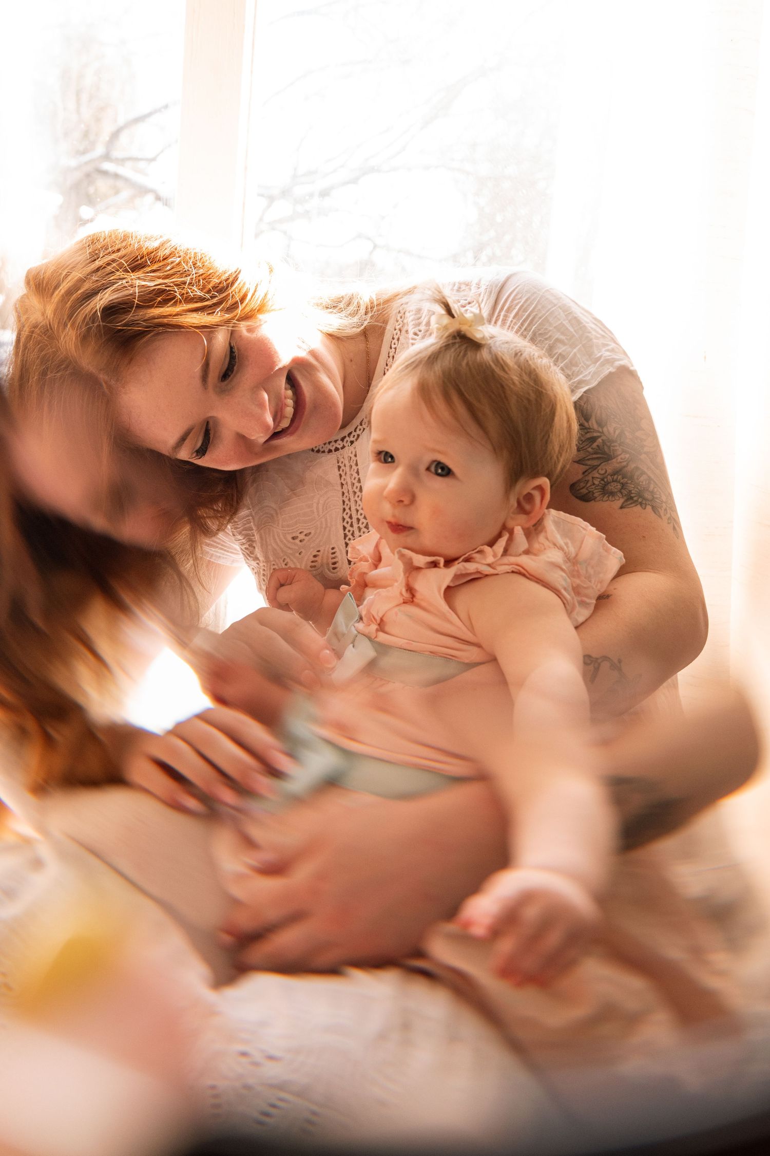 A smiling redhead mother holding her baby girl in soft natural light near a window.