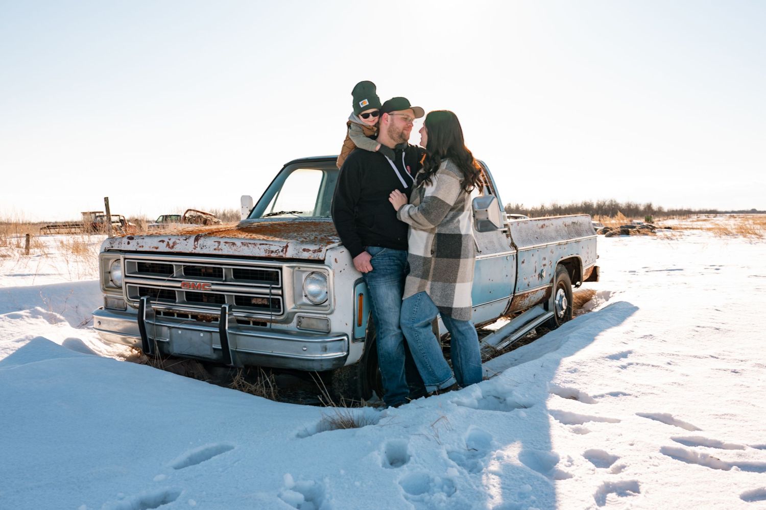 Couple standing together in snow beside a vintage rusty pickup truck on a sunny winter day.