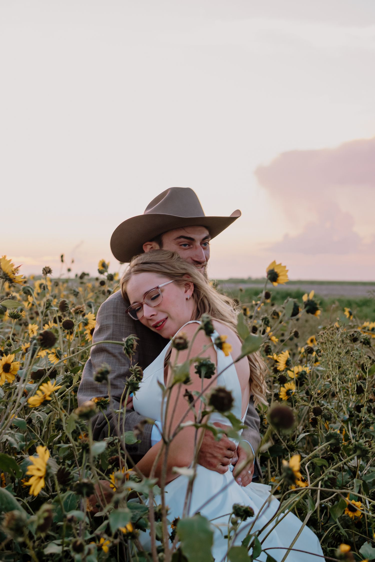 A person in a cowboy hat sits among blooming sunflowers during a golden sunset in a field.