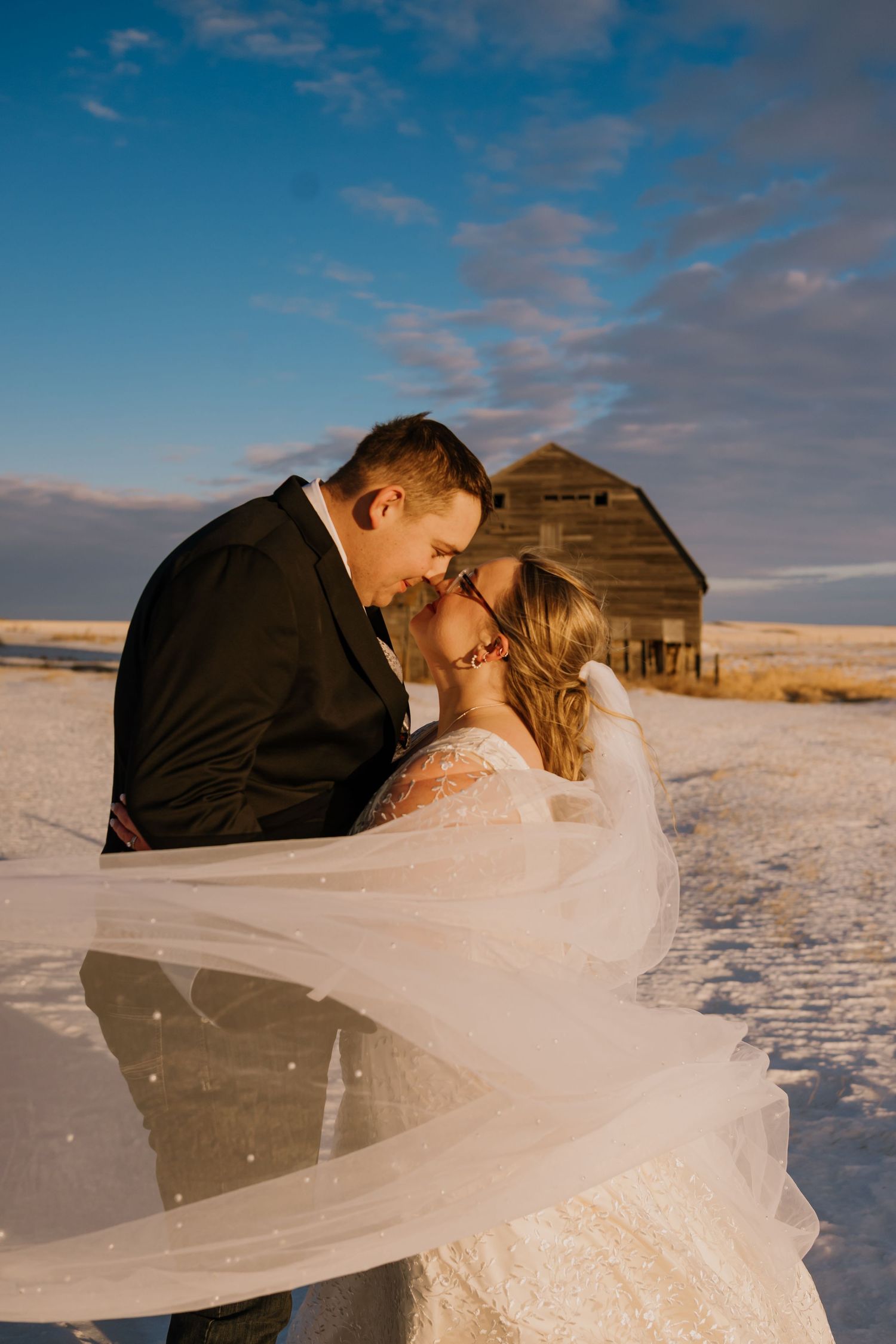 A romantic winter wedding photo with a couple kissing in front of a rustic barn during golden hour sunset.