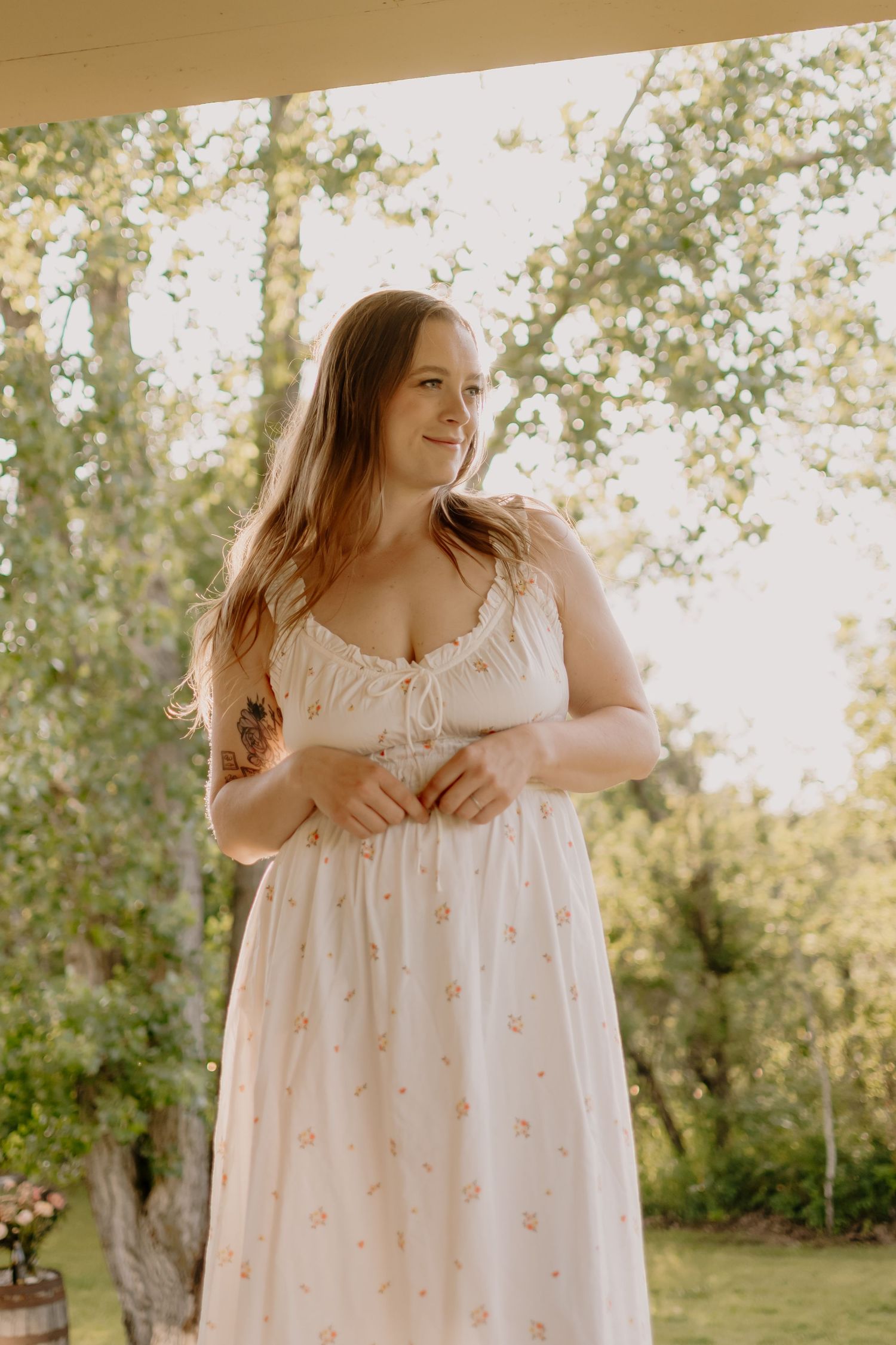 A person in a flowy white floral dress poses outdoors in a garden setting during golden hour lighting.