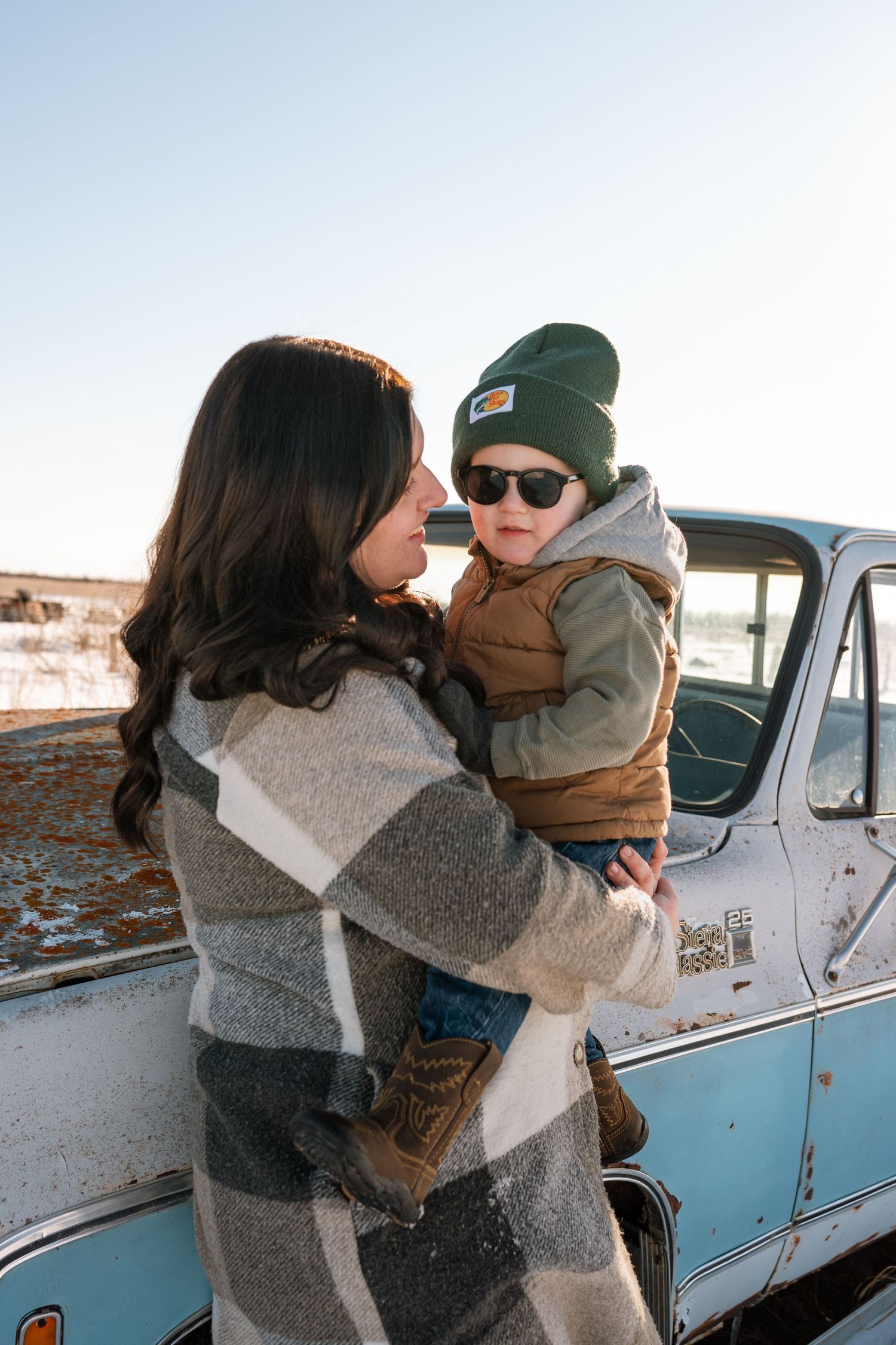 Mother in plaid coat holds young boy wearing sunglasses and green beanie by vintage truck.