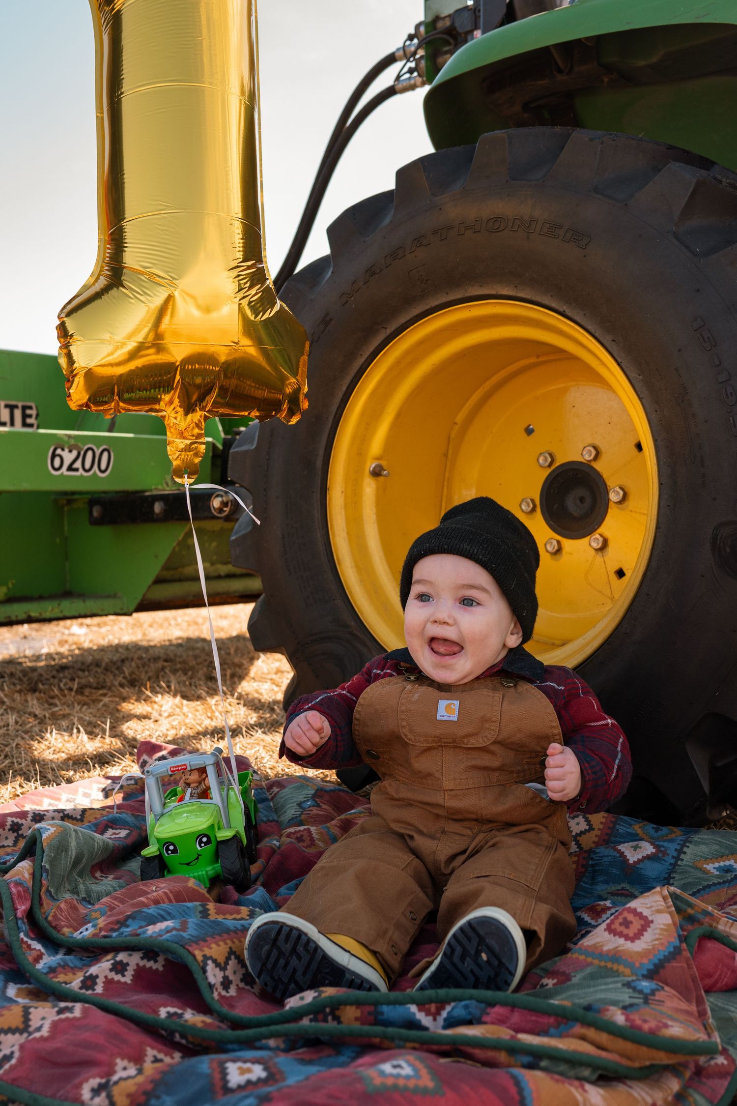 A smiling baby in Carhartt overalls sits beside a John Deere 6200 tractor tire with a gold number one balloon on their first birthday.