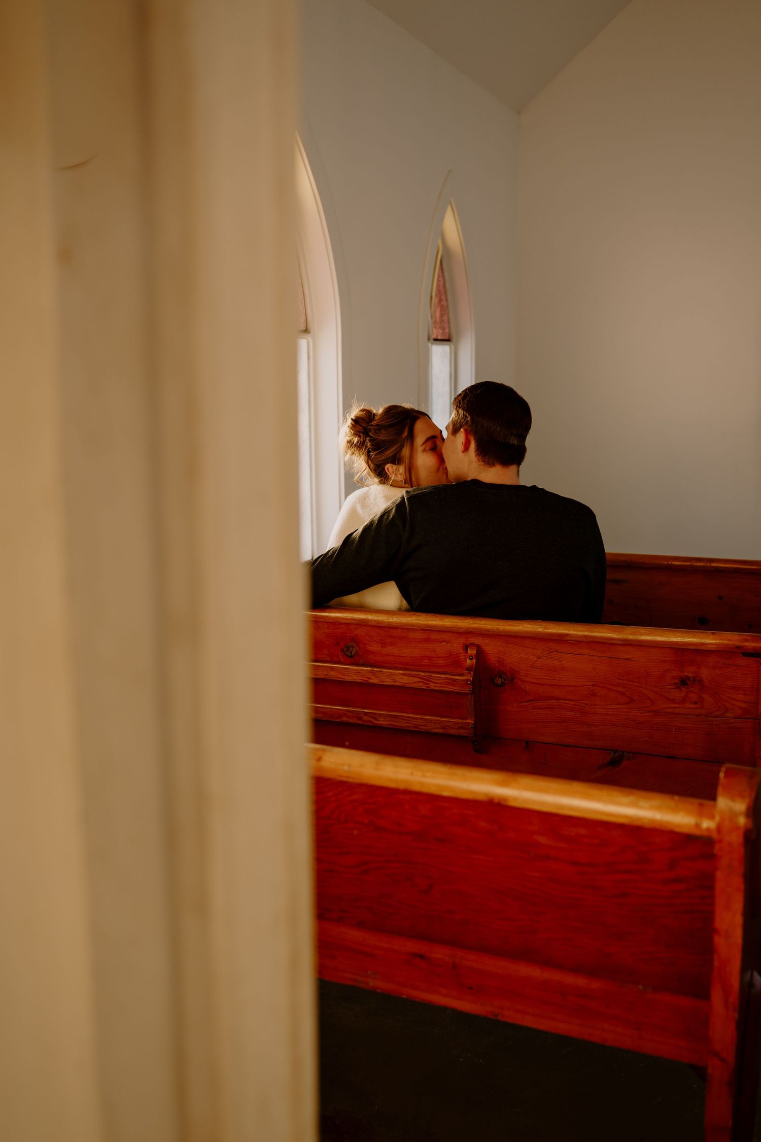 Two people sit in wooden church pews with sunlight streaming through windows in a serene interior space.