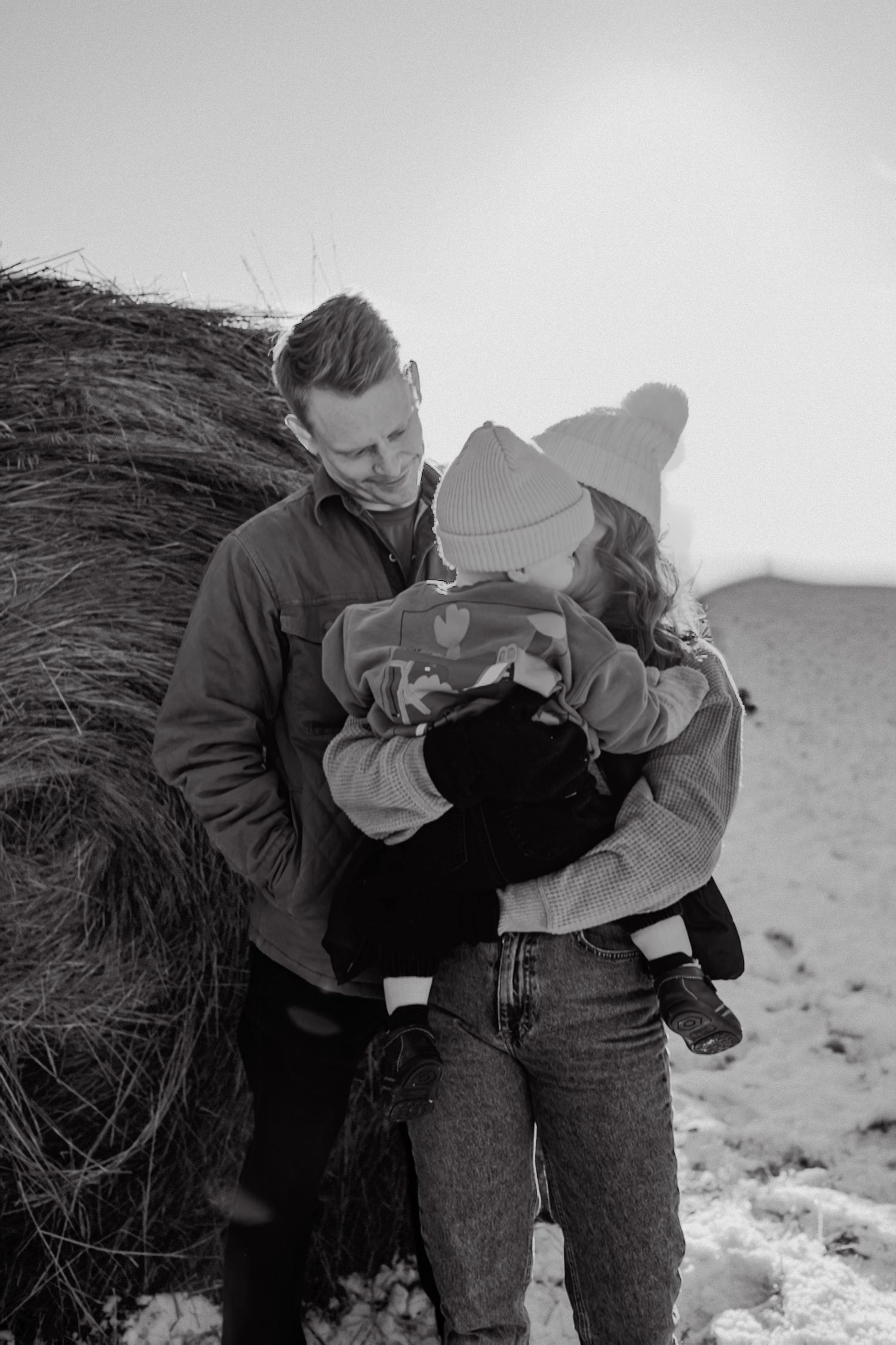 Two figures in dark coats stand close together in a winter landscape with hay bales visible in the background.