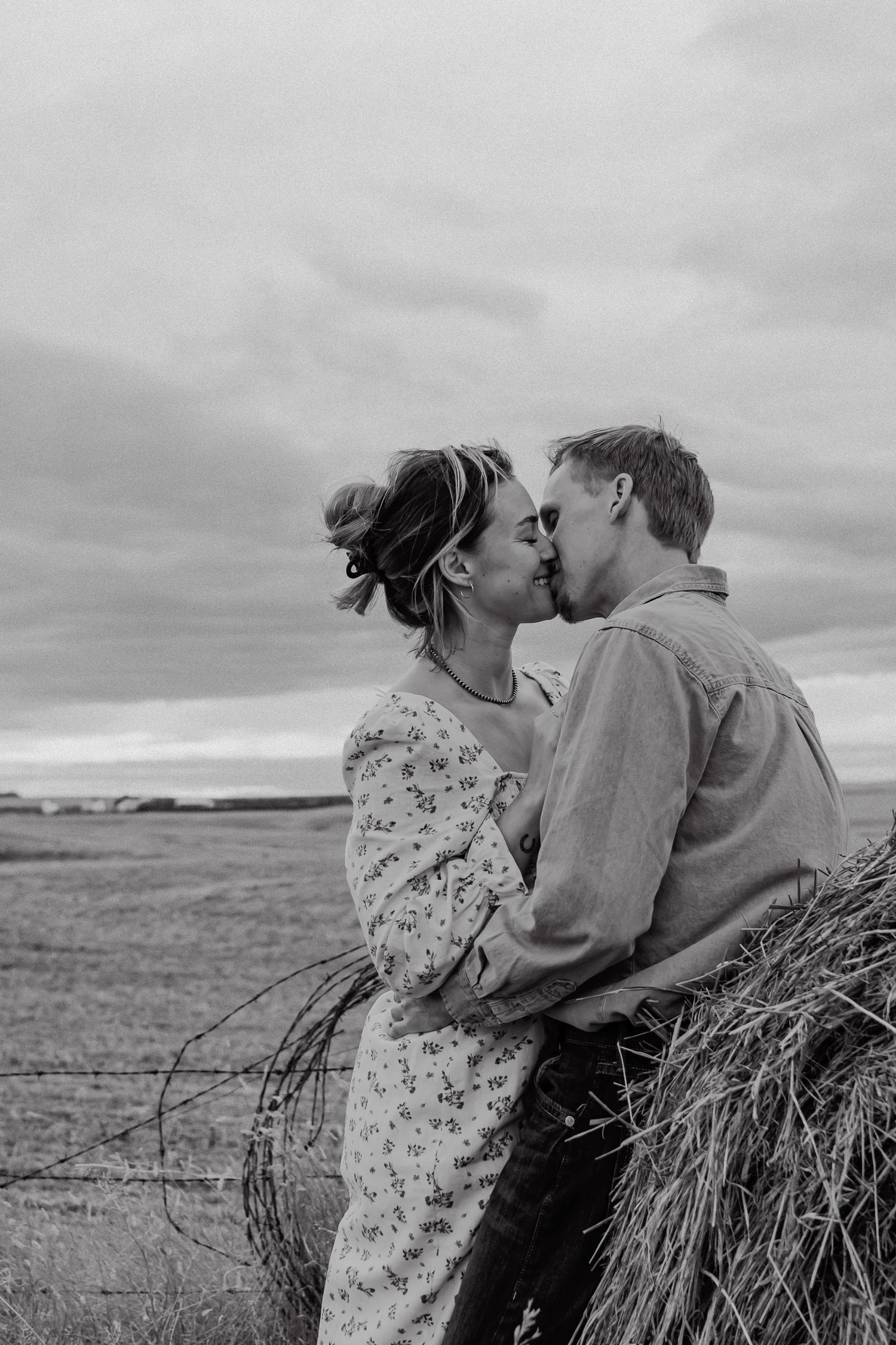 A romantic black and white photograph of a couple embracing near a hay bale in a rural field.
