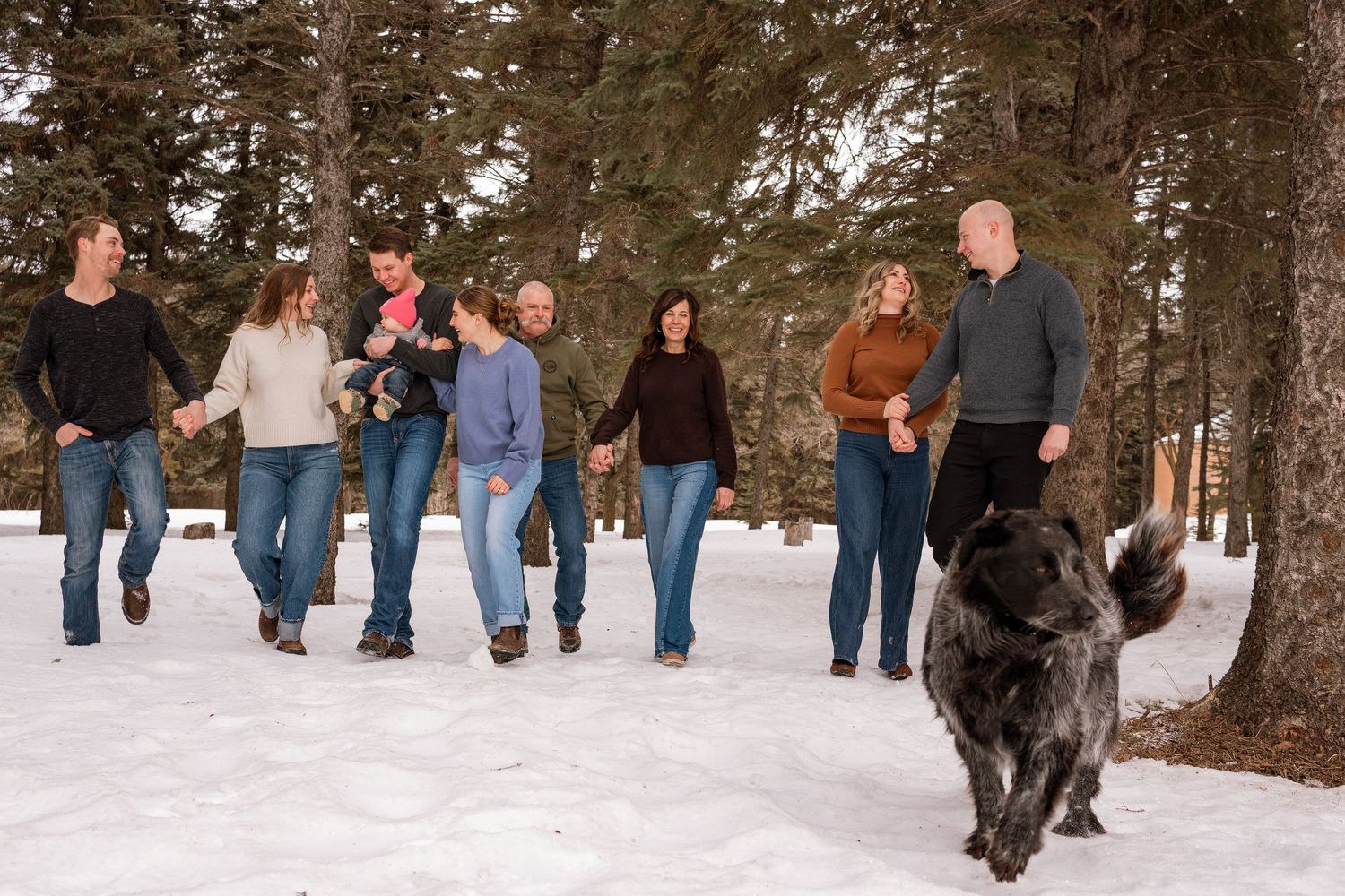 Family of seven walks hand-in-hand in snowy forest with large black dog in foreground during winter.