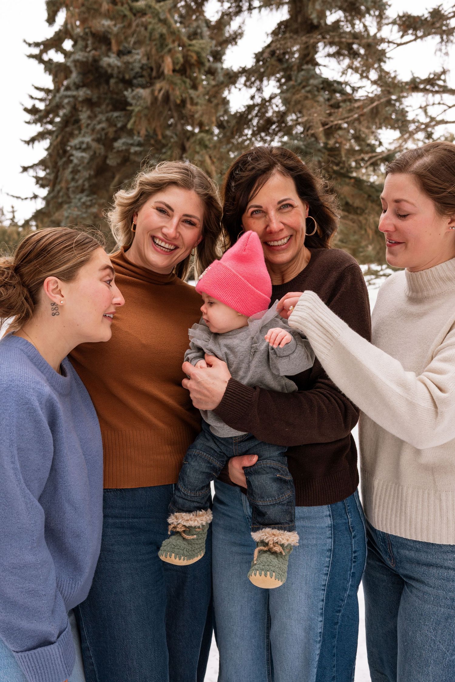 Four women smile and laugh together outdoors in winter, admiring a baby in a pink hat amid snowy trees.