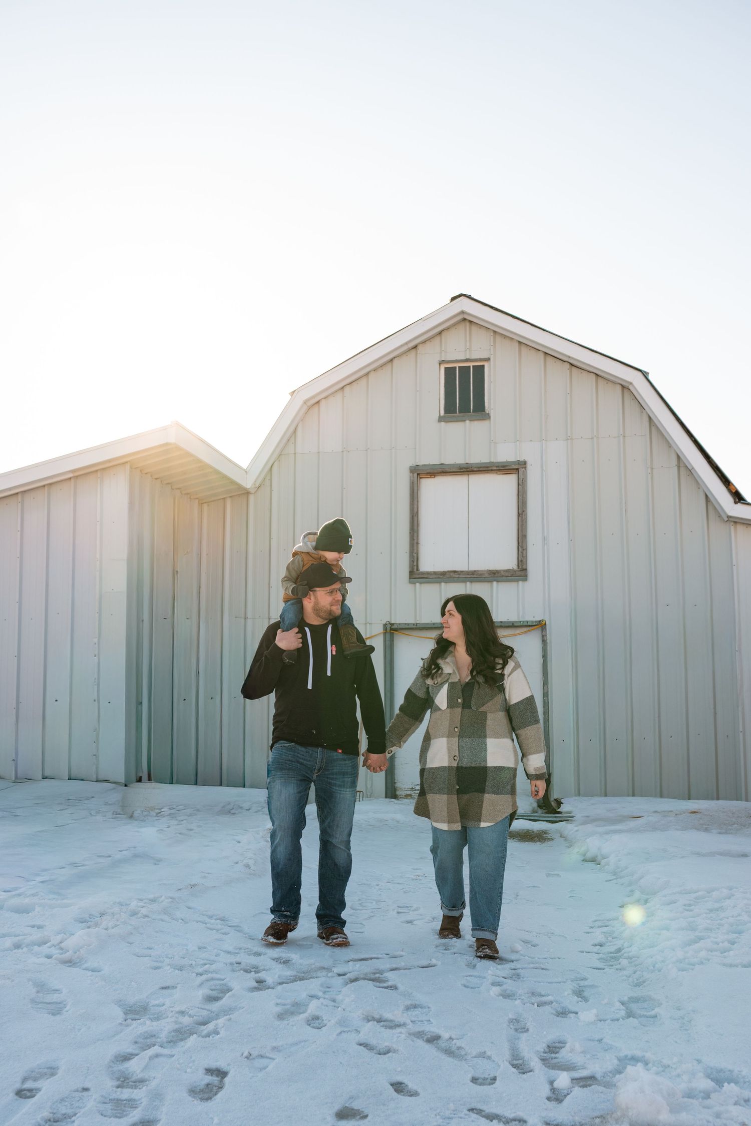 Couple holds hands walking in snow in front of white barn on sunny winter day.