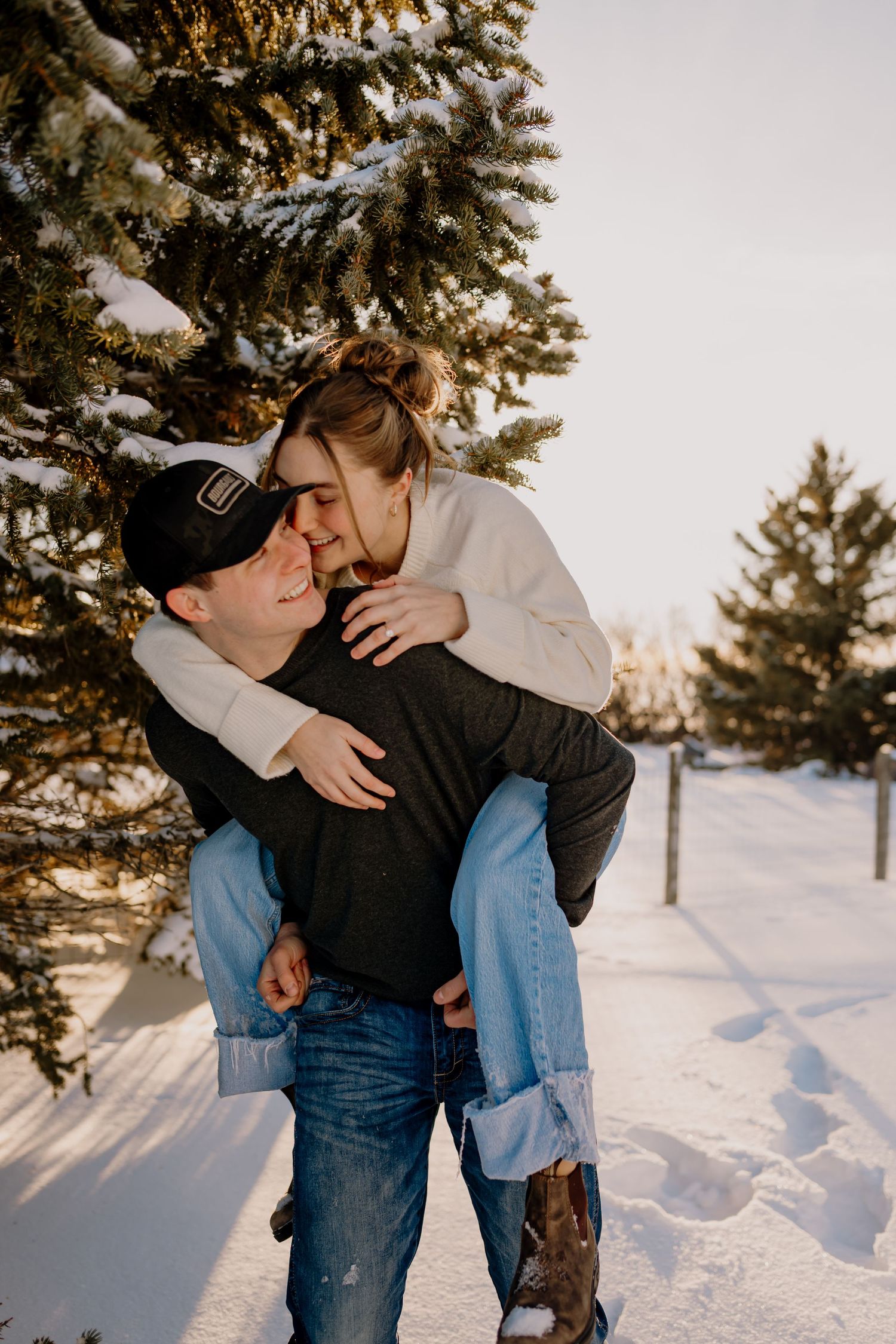 A couple shares a tender piggyback moment in a snowy winter landscape surrounded by evergreen trees at sunset.