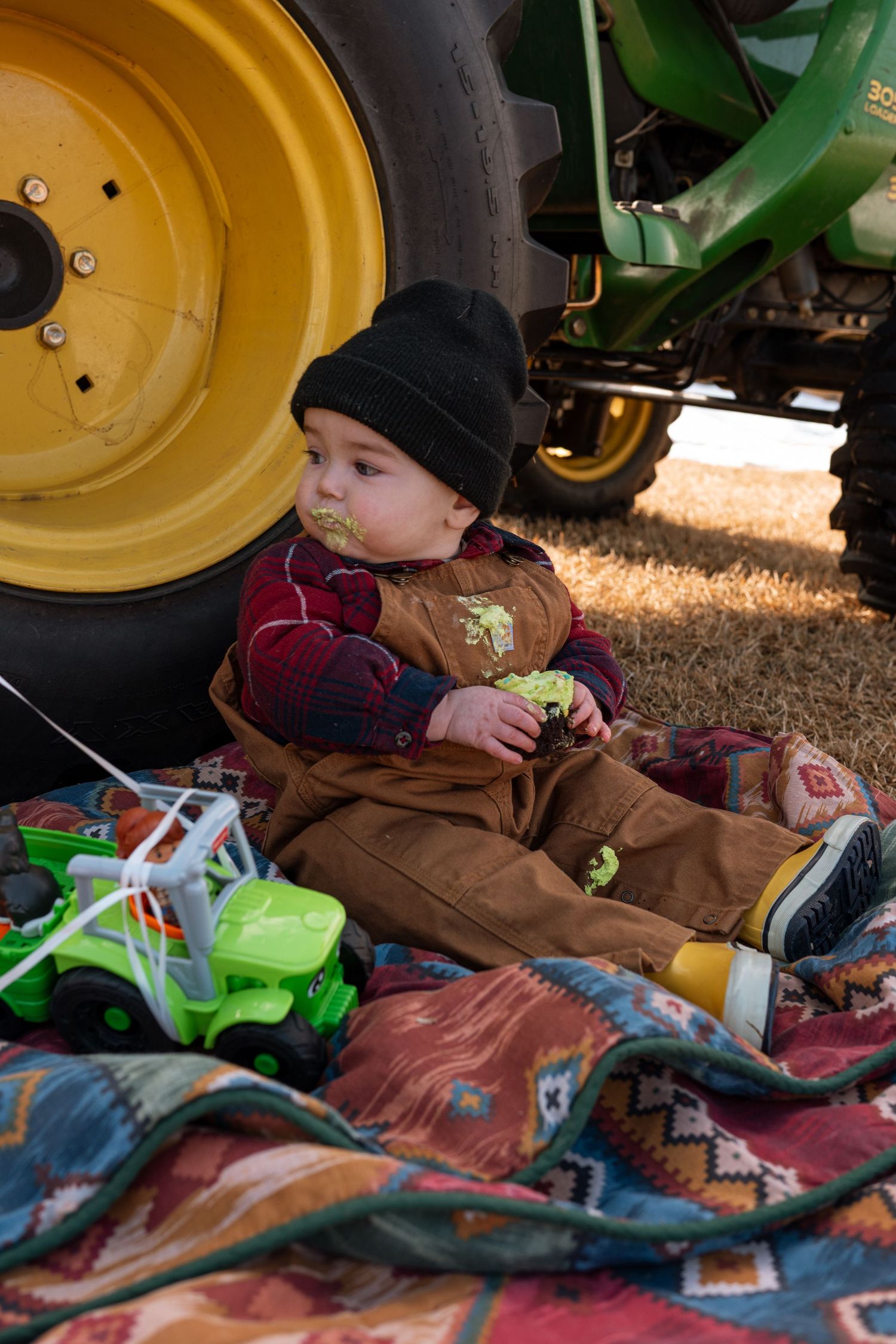 Baby in overalls and black beanie sits on blanket beside John Deere tractor tires, playing with toy trucks outdoors.