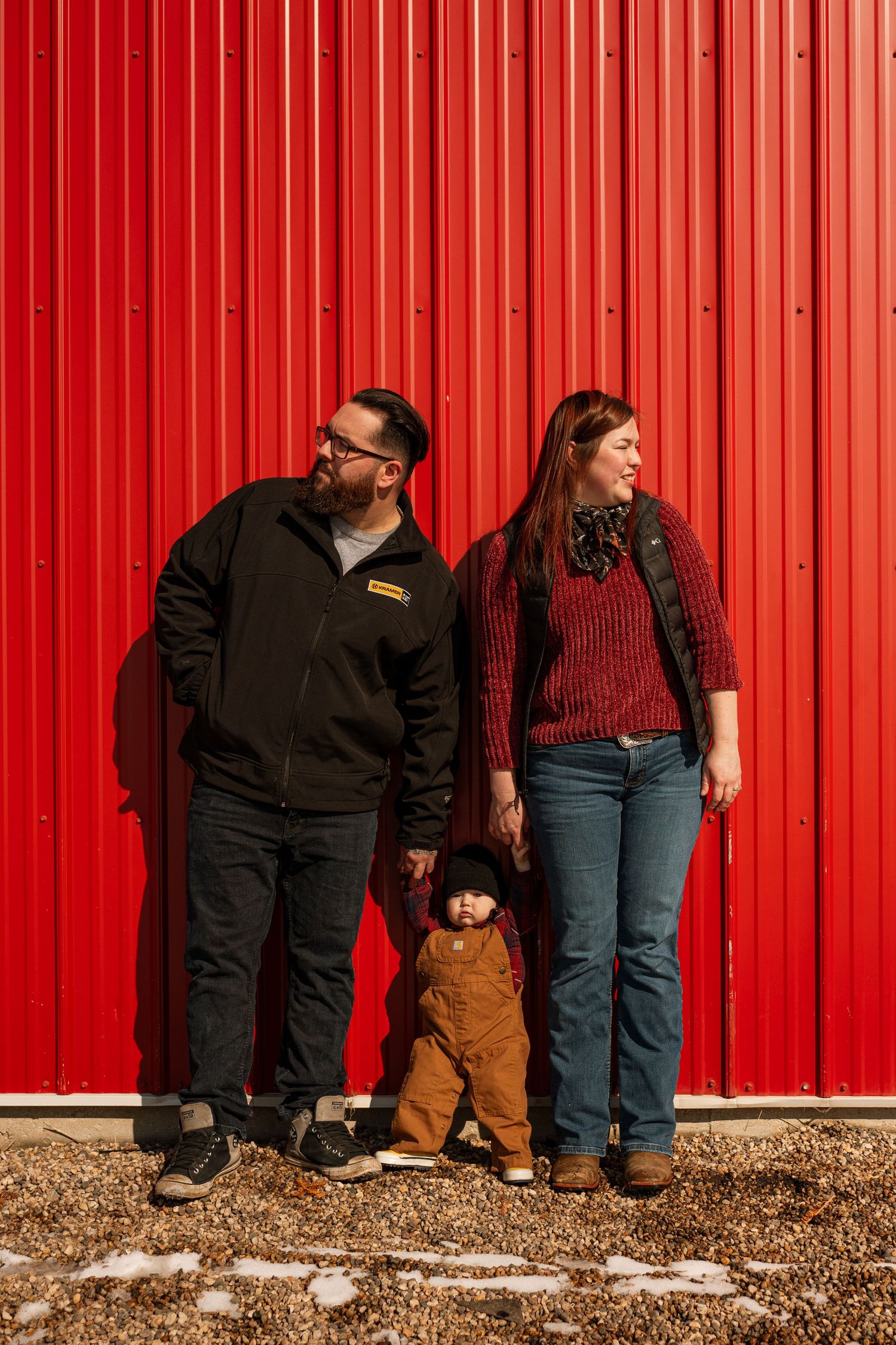 Family of three posing in front of a bright red metal wall, parents holding toddler's hands outdoors.